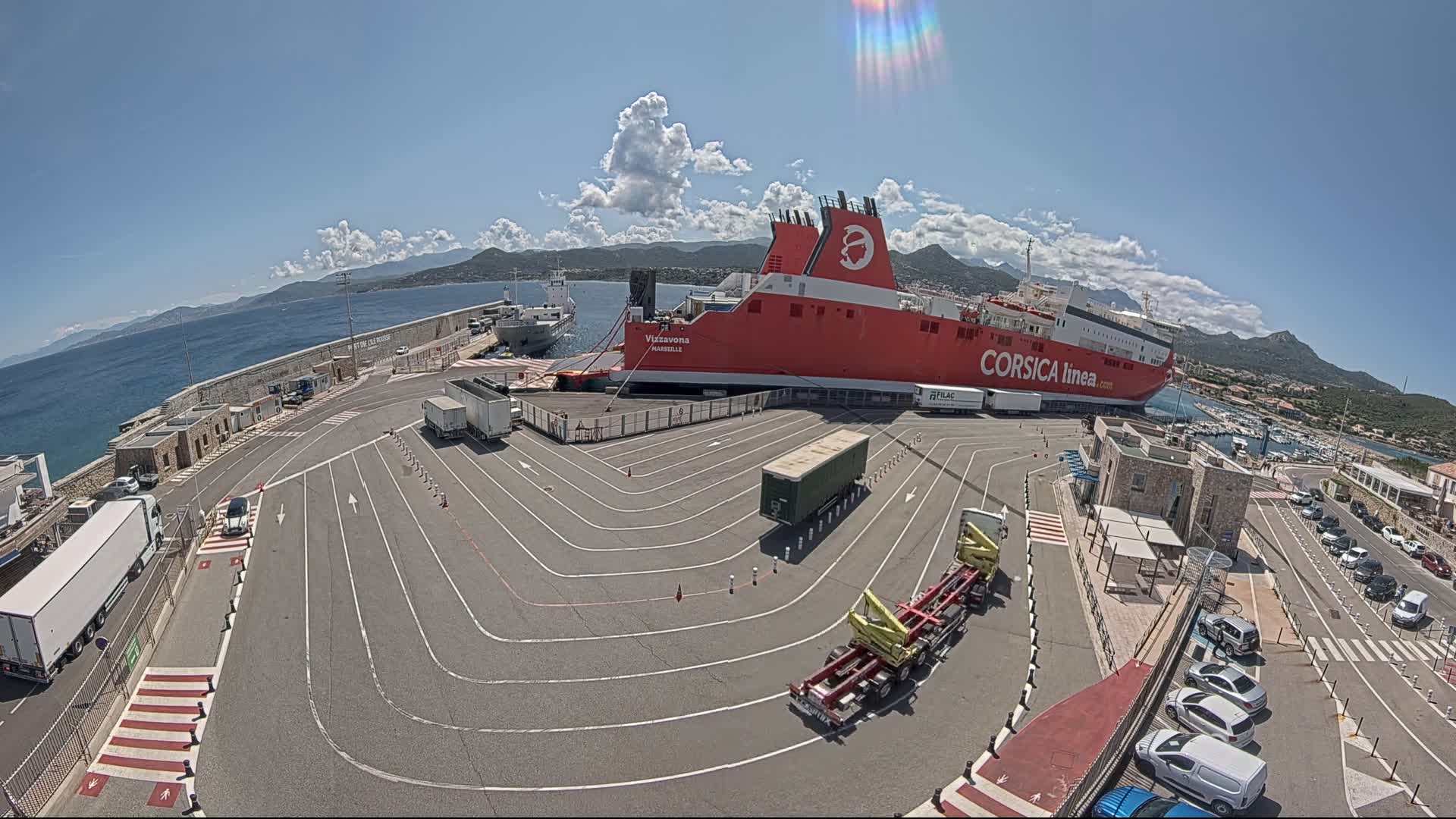 A large red ferry is docked at a port on a sunny day with a few clouds, surrounded by trucks, cars, and a roadway marked with lane dividers.