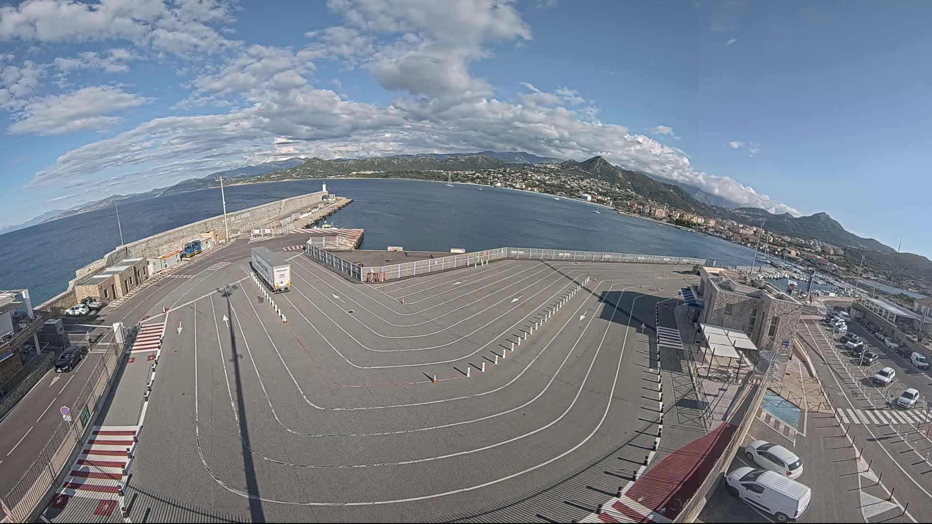 A mostly empty ferry terminal with marked lanes, overlooks a calm bay under a partly cloudy sky, mountains visible in the distance.