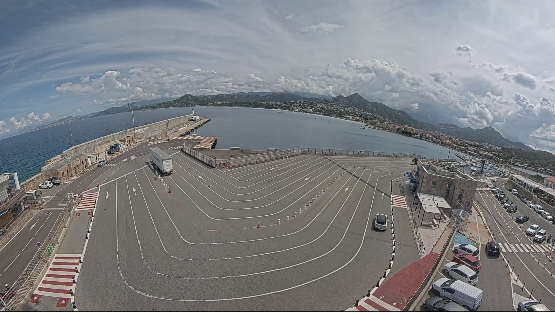 A ferry terminal with a large, marked parking area overlooks a calm bay under a partly cloudy sky.
