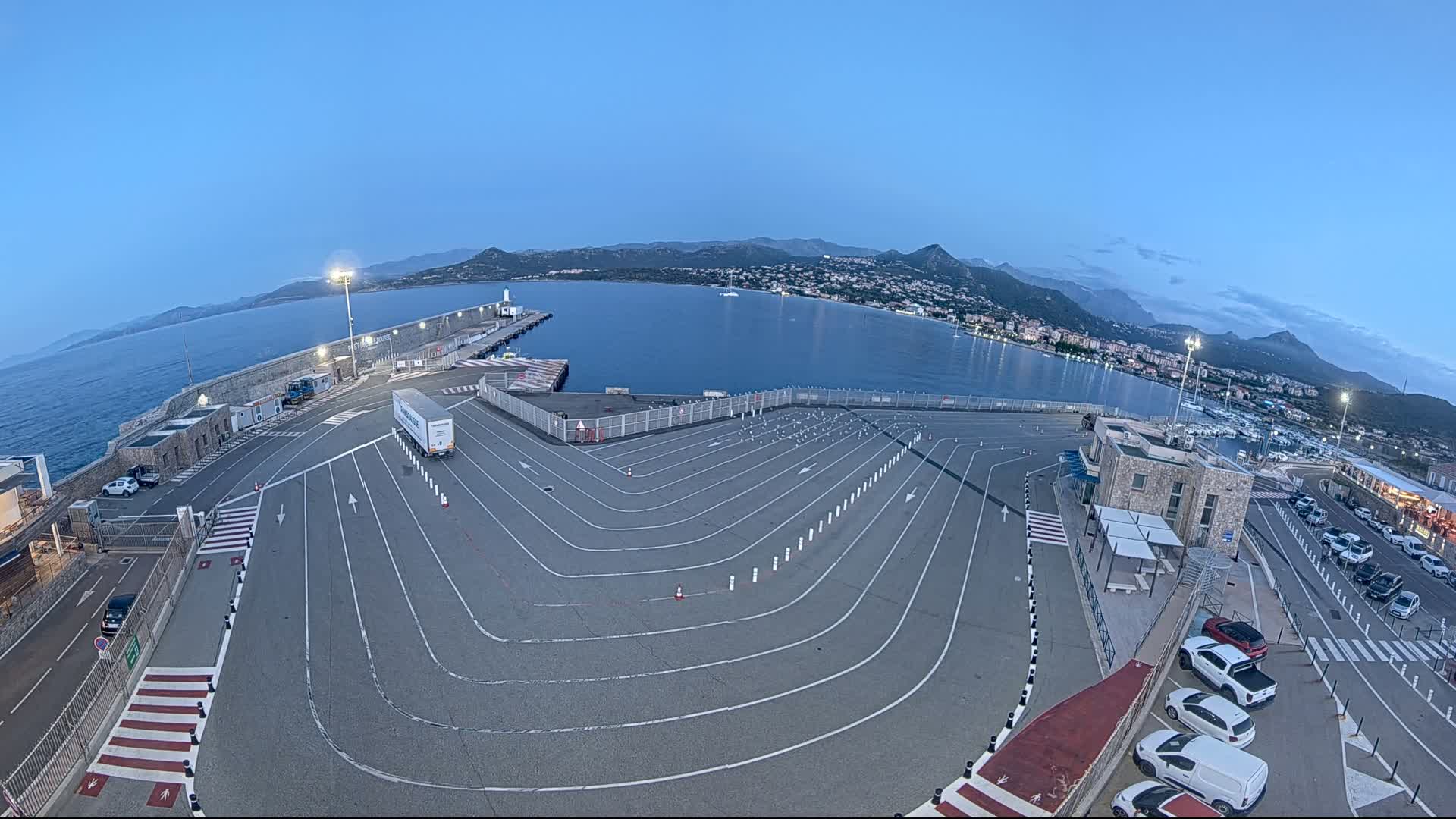 An empty ferry terminal with marked lanes overlooks a calm bay and distant mountains under a clear twilight sky.