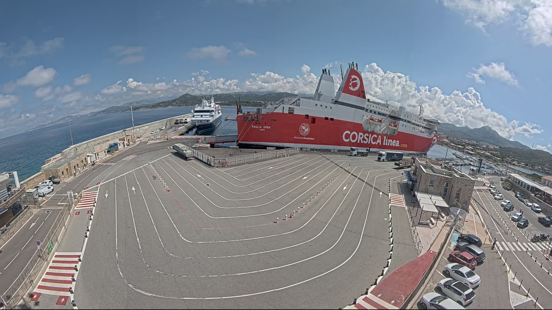 A large red ferry is docked at a port on a sunny day, with another smaller ferry visible in the background, and cars parked in a nearby lot.