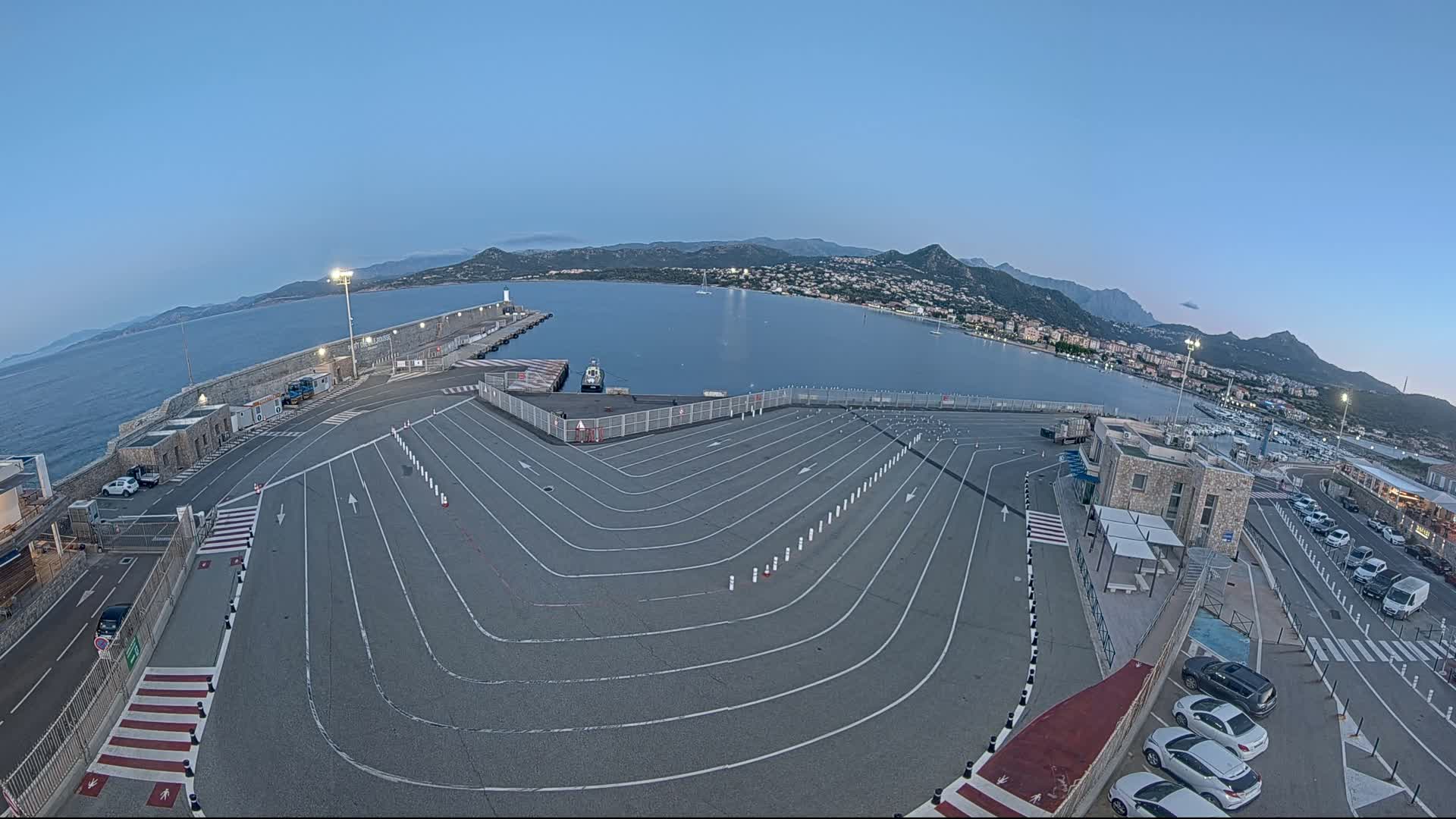An empty ferry terminal with marked parking spaces overlooks a calm bay and mountainous coastline under a clear, twilight sky.