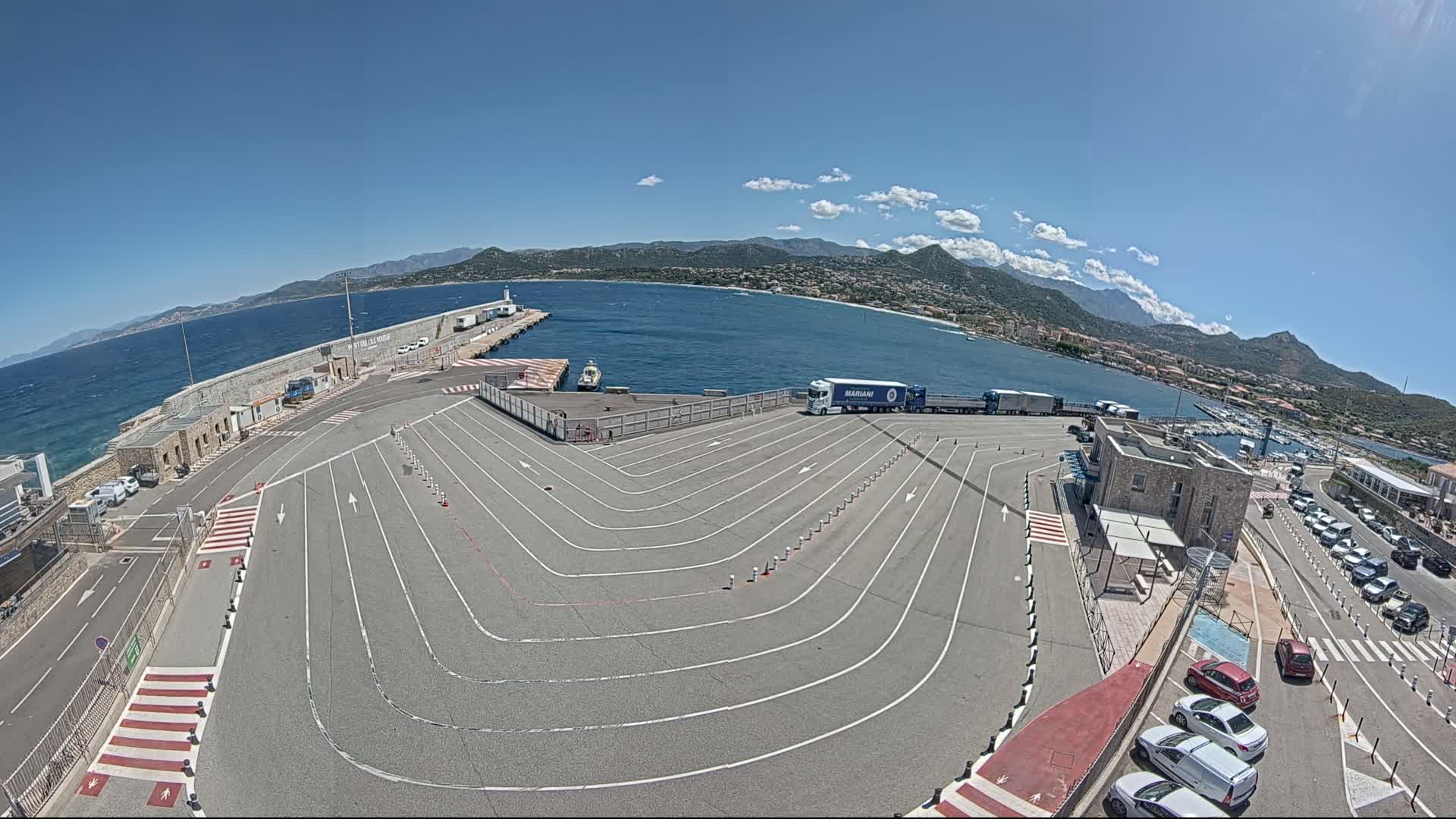 A ferry terminal with a large, curving parking area overlooks a calm sea and mountains under a clear blue sky.