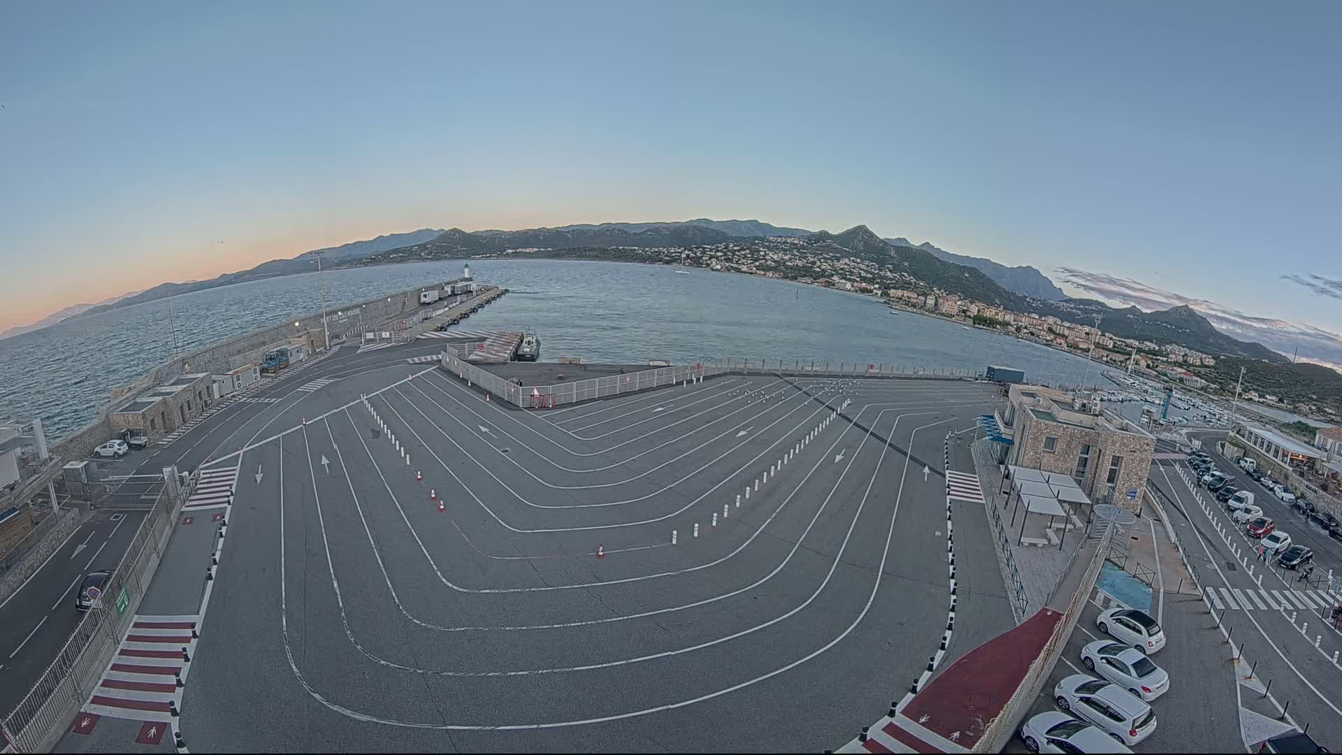 An empty ferry terminal with marked lanes curves toward a calm body of water under a clear, twilight sky, mountains visible in the distance.