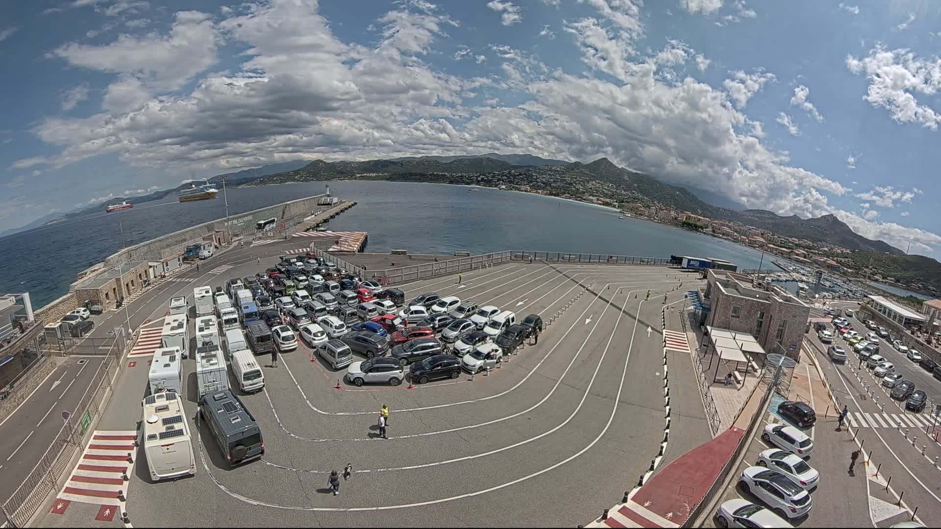 A ferry terminal with numerous cars and camper vans parked in designated lanes, overlooking a calm bay under a partly cloudy sky.