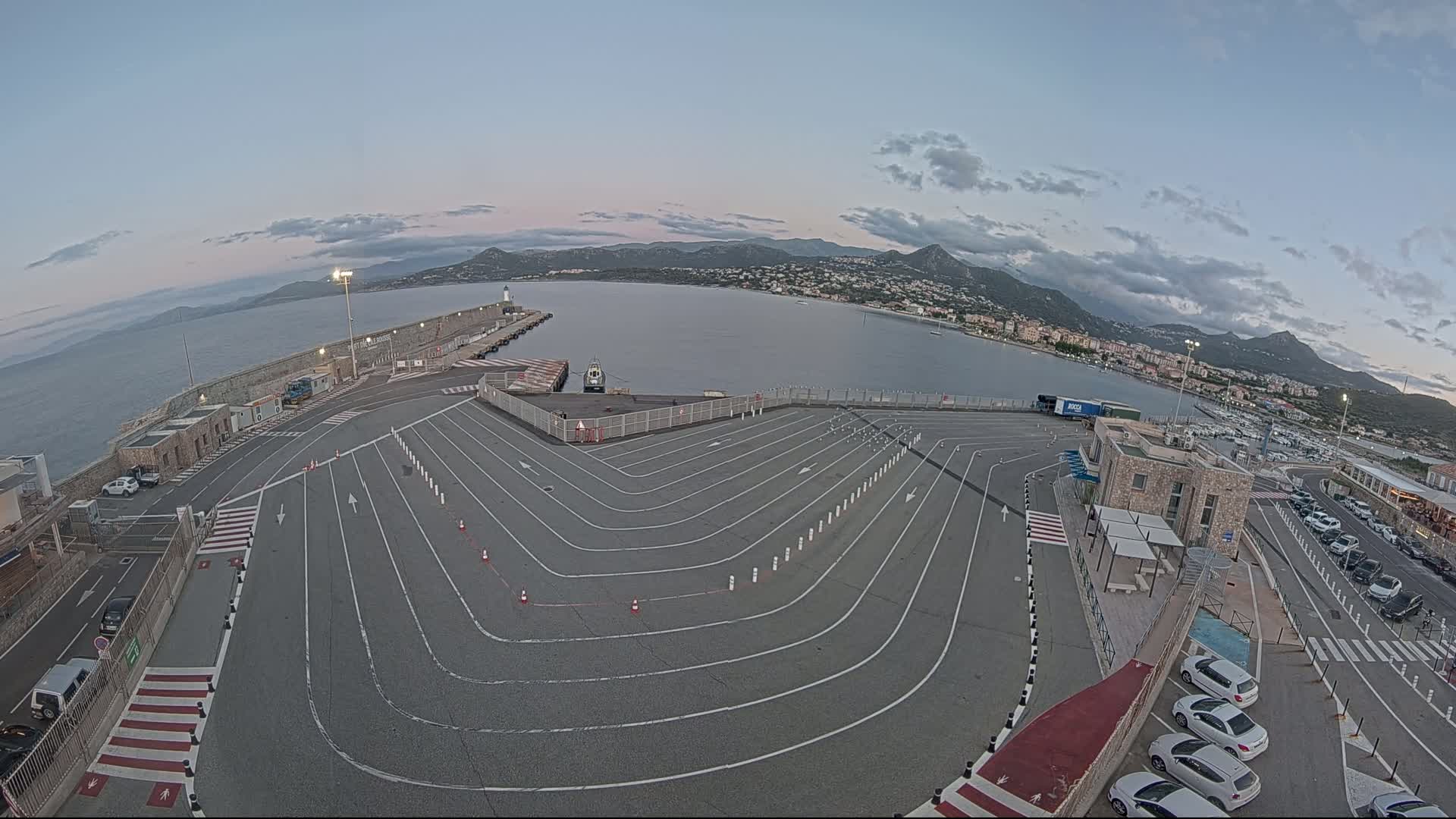 An empty ferry terminal with marked lanes curves around a harbor under a partly cloudy twilight sky, overlooking a town nestled against mountains.
