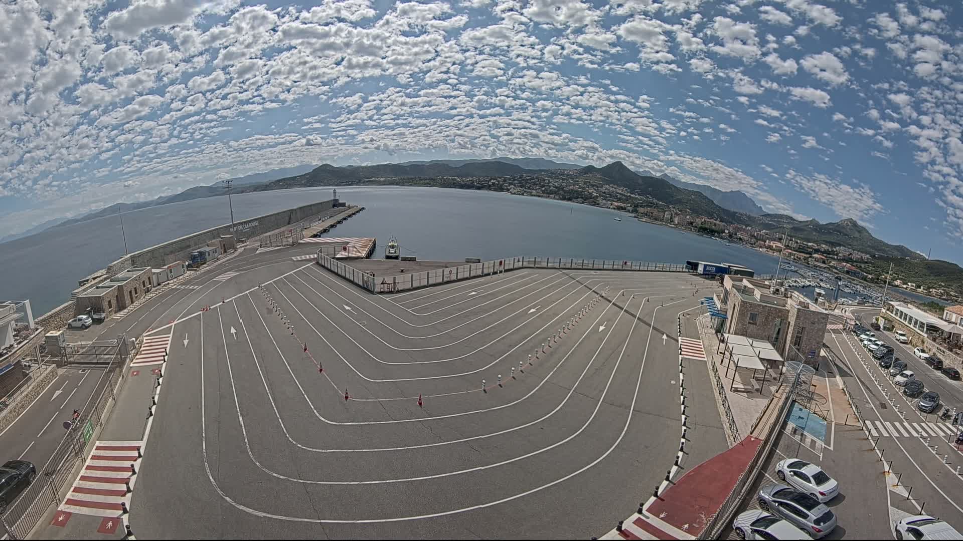 An aerial view shows a ferry terminal with a large, marked parking area, a calm body of water with mountains in the background, under a mostly sunny sky with scattered clouds.