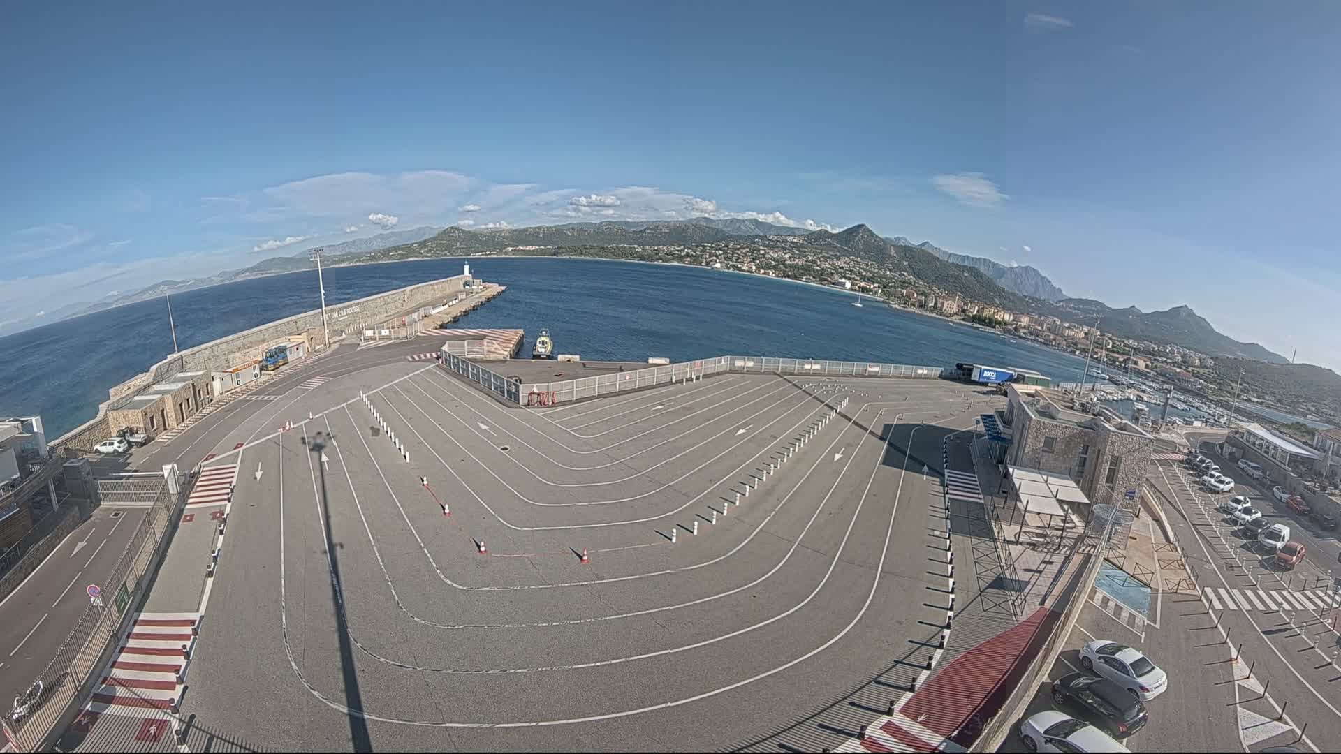 An empty ferry terminal parking lot with marked lanes curves around a small building, overlooking a calm blue sea and distant mountains under a clear blue sky.