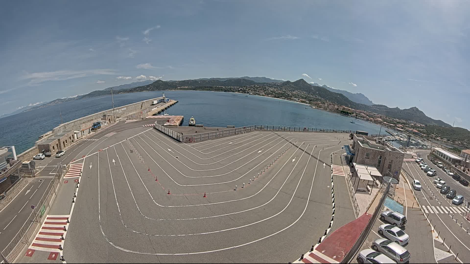 An aerial view of a ferry terminal with a large, marked queuing area for vehicles, under sunny skies, overlooking a calm bay and mountainous coastline.