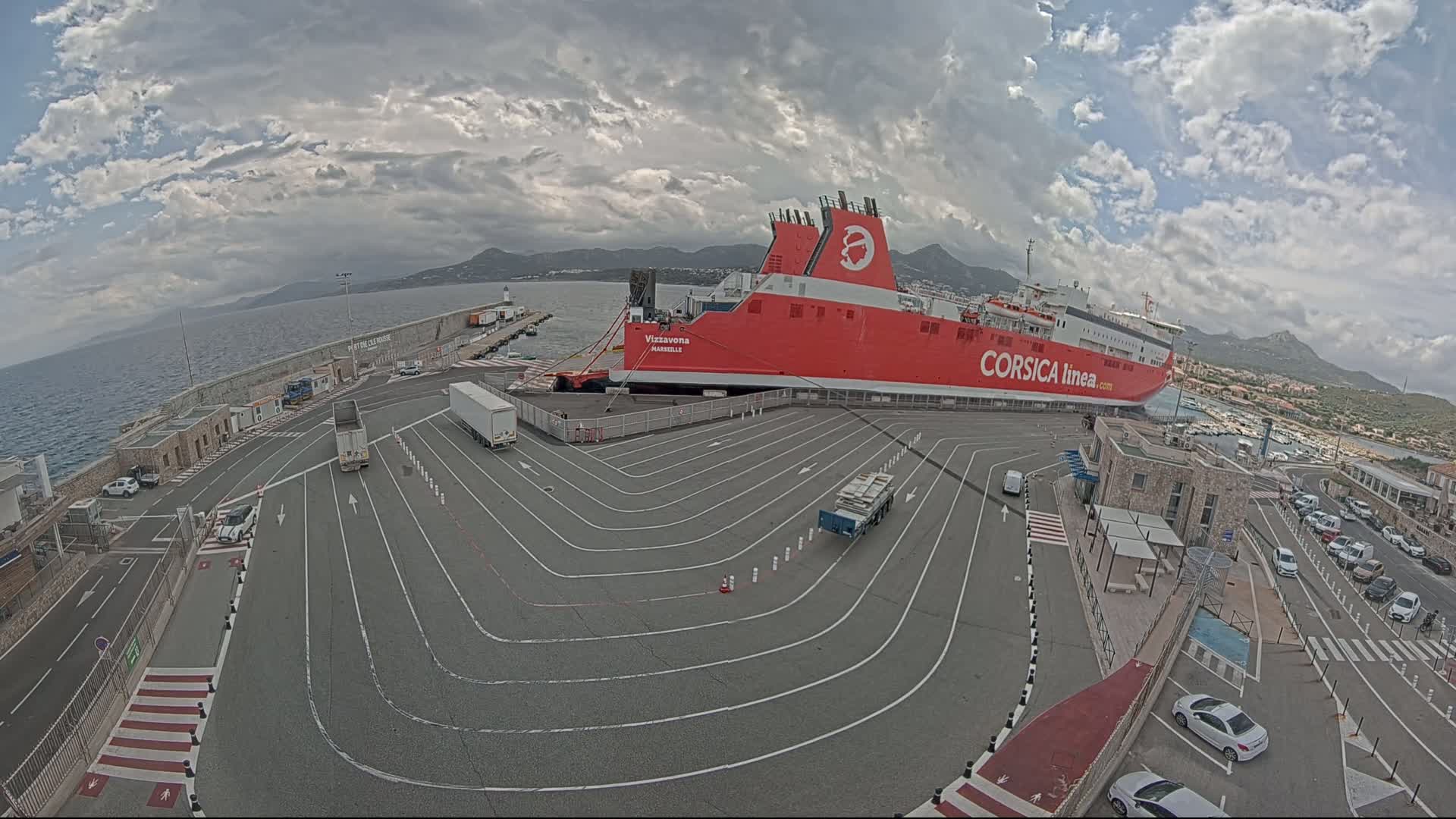 A large red ferry is docked at a port under a partly cloudy sky, with several vehicles parked in the surrounding area.