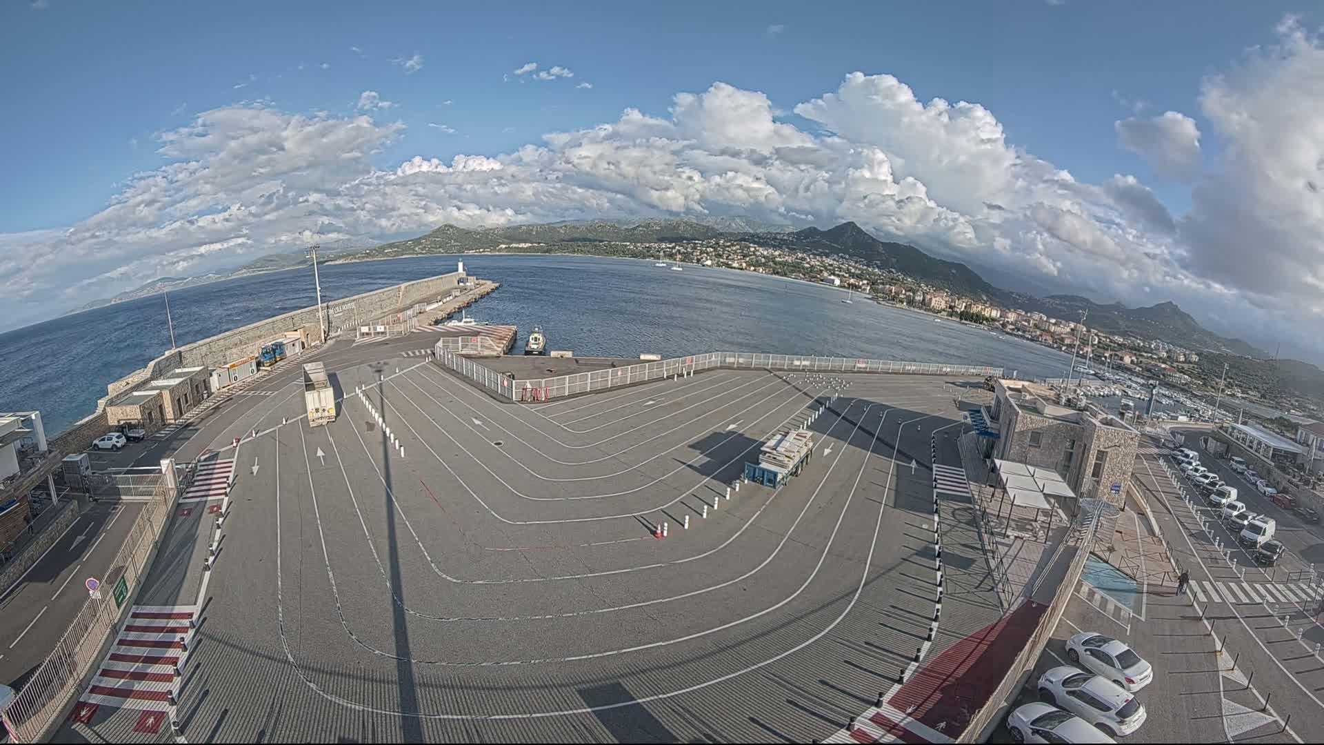 A mostly sunny day at a ferry terminal with a large paved area, marked lanes, and several vehicles parked, overlooking a calm body of water and distant mountains.