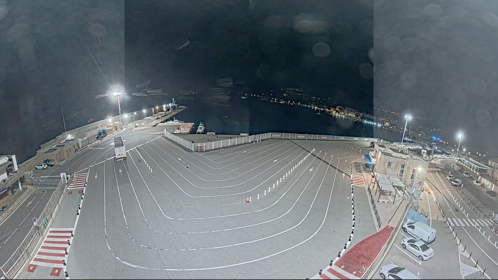 An empty, paved ferry terminal at night is shown under a dark, cloudy sky, with a few vehicles parked nearby and a body of water visible in the distance.