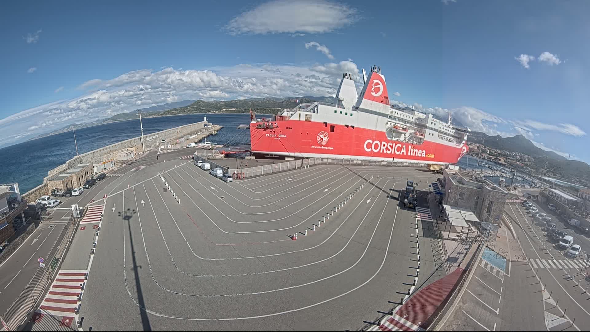 A large red and white ferry is docked at a port under a partly cloudy sky, with cars parked in designated lanes nearby.