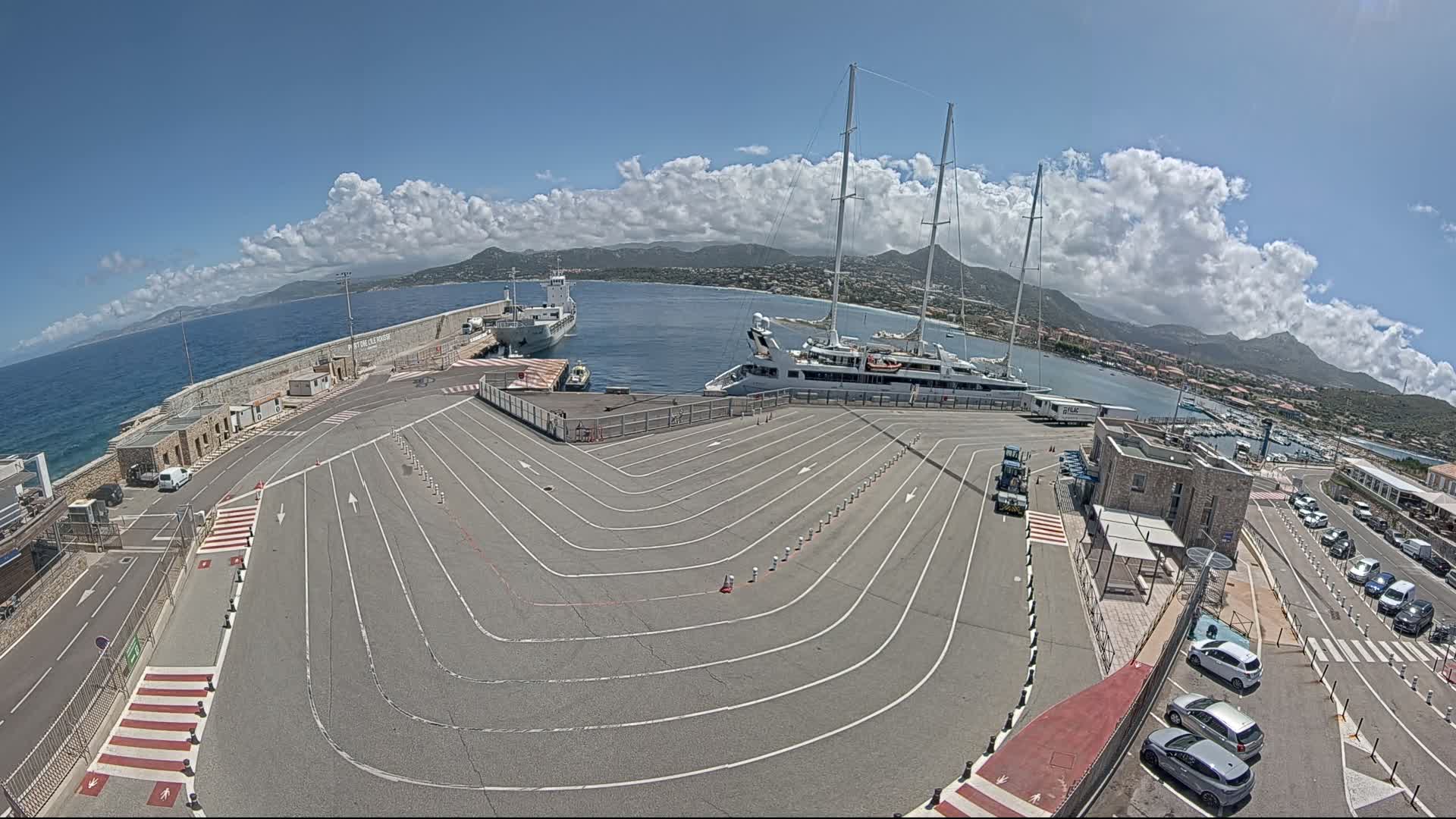 A harbor with a large yacht and other vessels moored, under a partly cloudy sky, features a paved area with marked lanes leading to a dock.