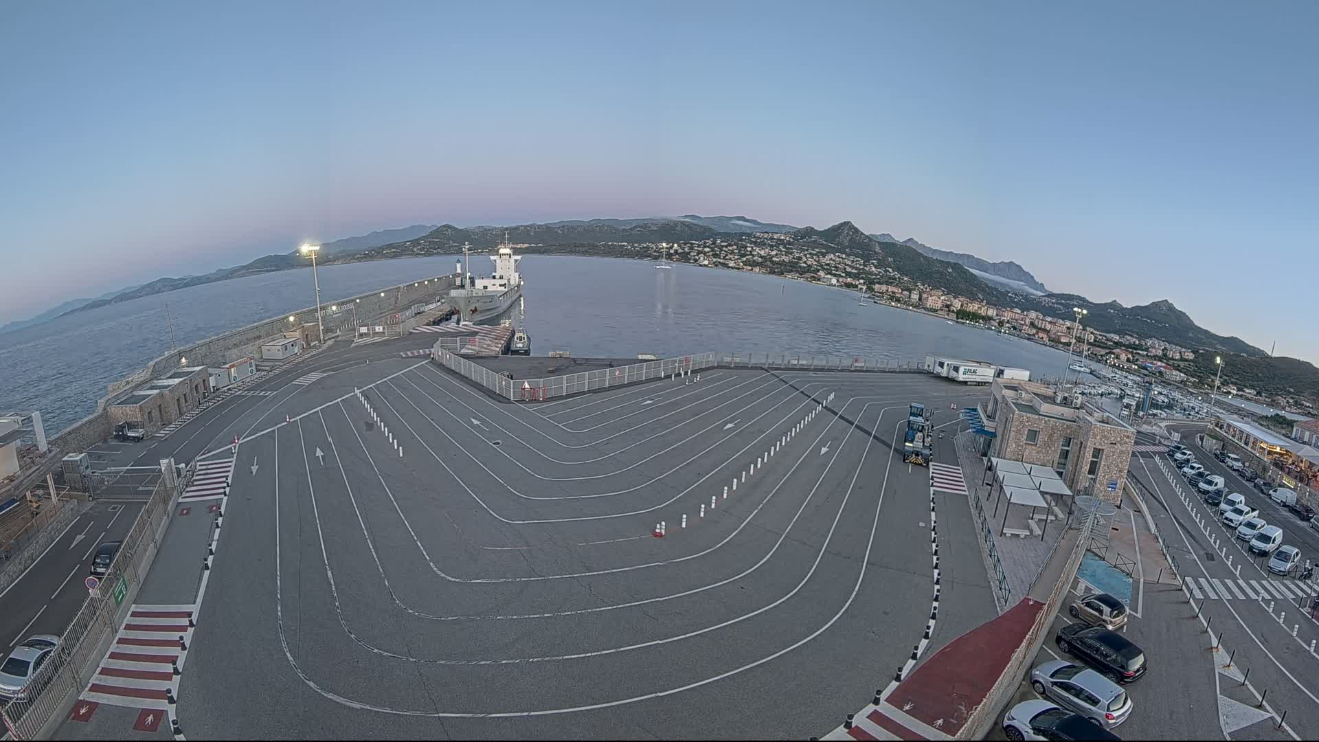 A ferry terminal with a large, curved parking area, a ship docked at the pier, and a calm body of water under a clear, twilight sky, backed by mountains.