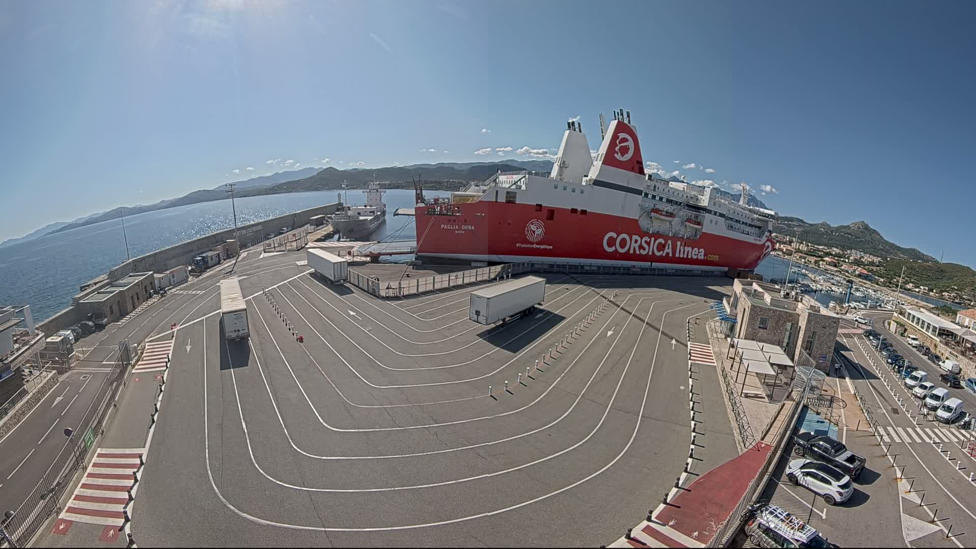 A large red and white ferry is docked at a port on a sunny day, with several trucks and cars parked nearby.