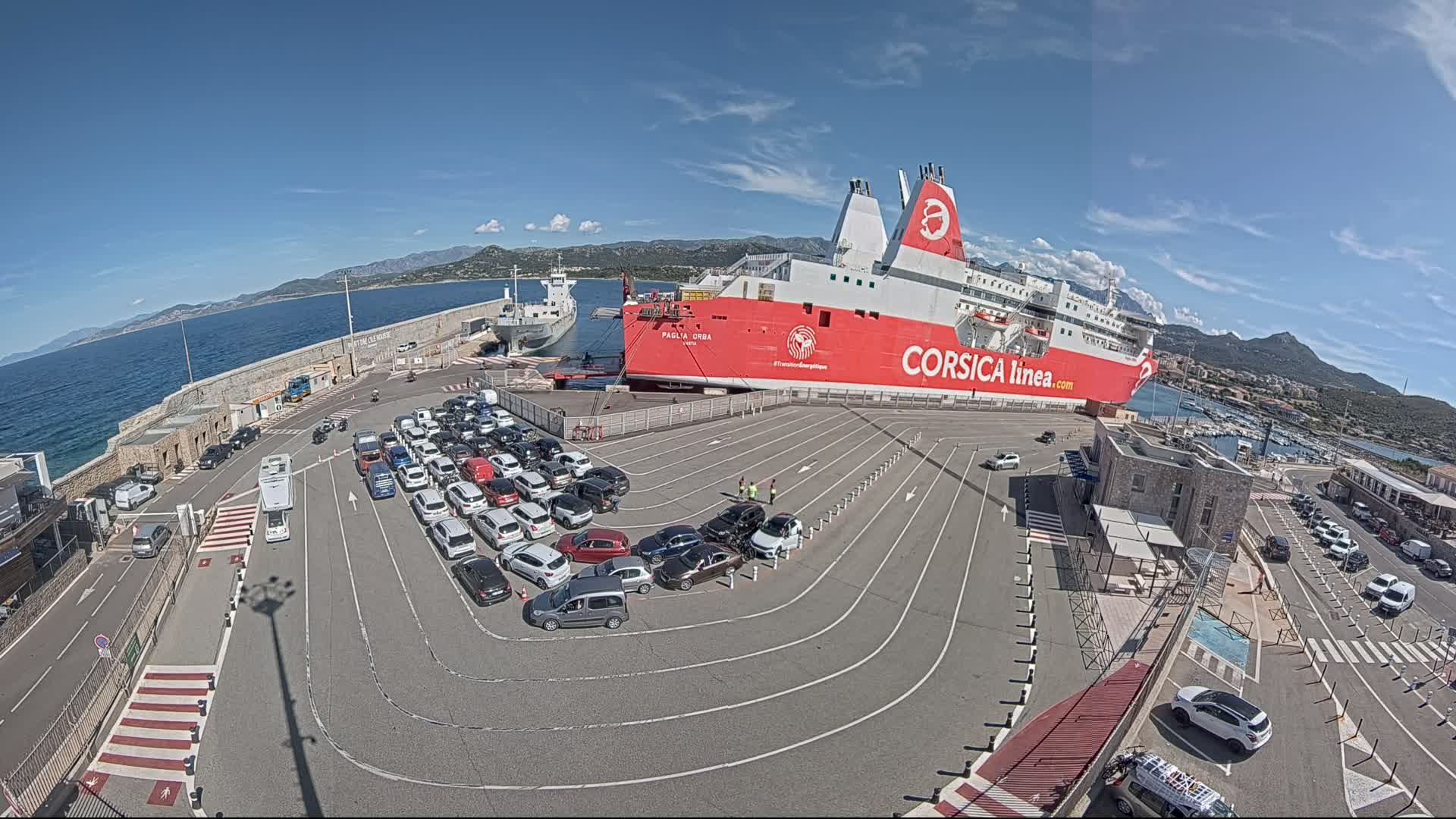 A large red and white ferry is docked at a port on a sunny day, with numerous cars parked in designated lanes nearby.