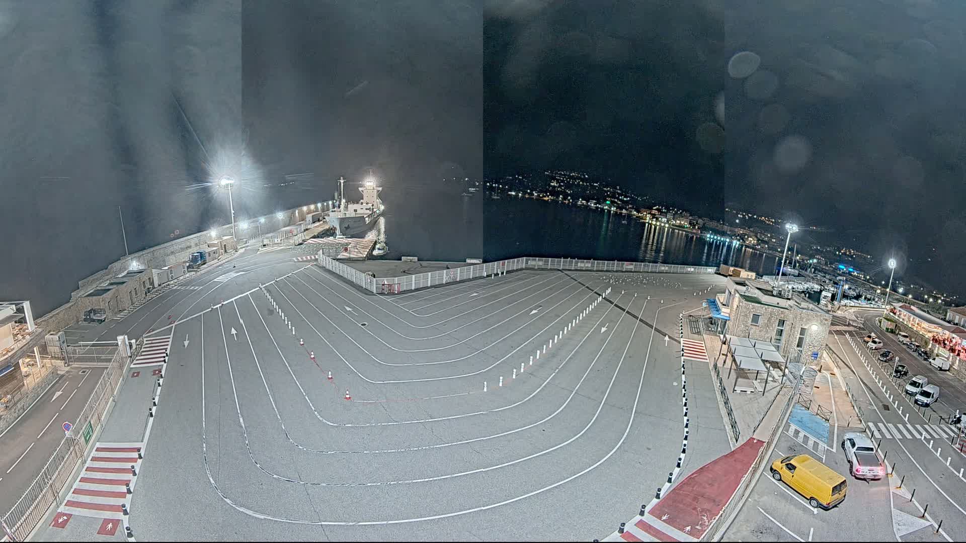 A nighttime aerial view of a mostly empty port parking lot with a ship docked at the water's edge under a dark, cloudy sky.