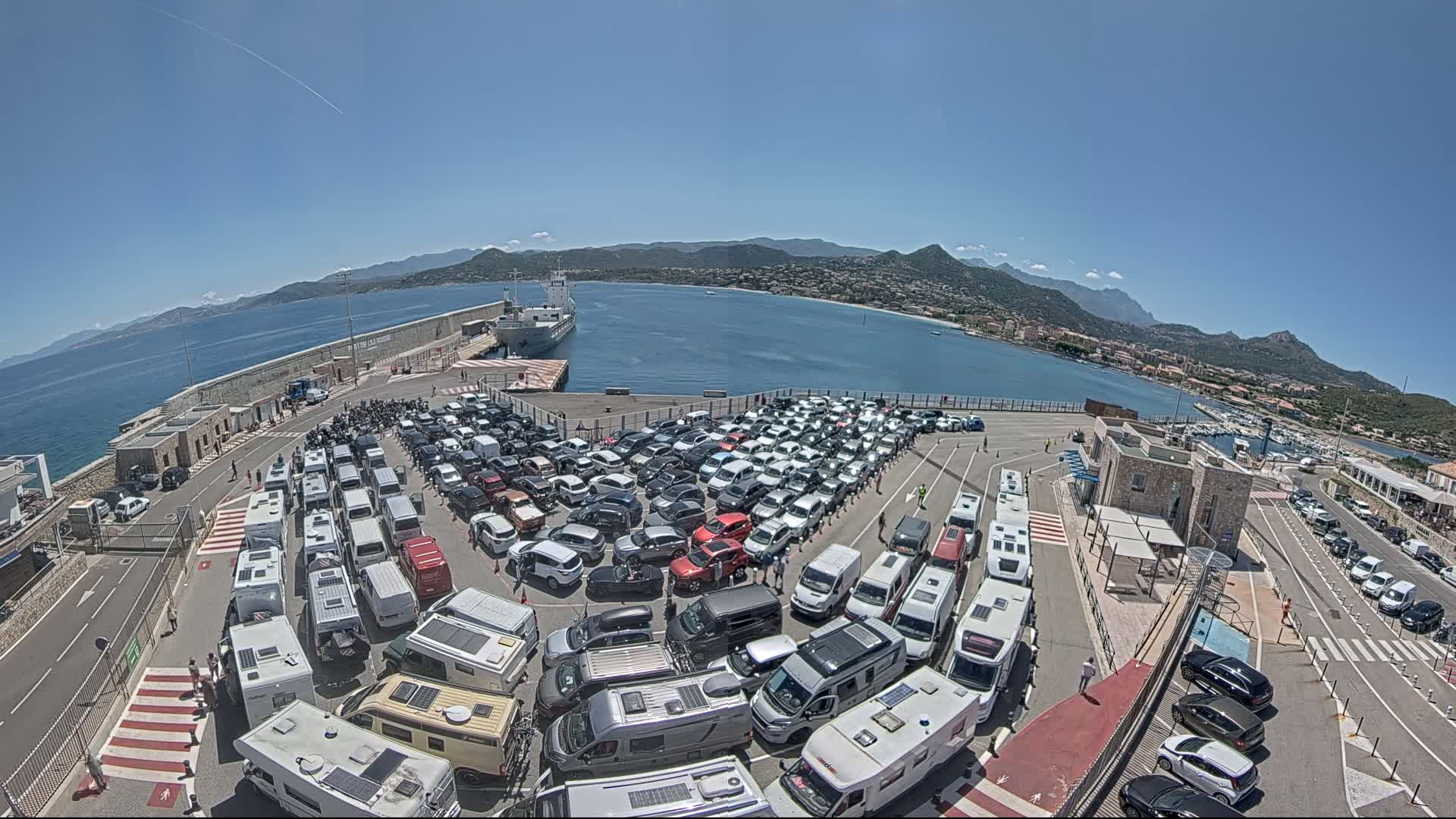 A large ferry terminal on a sunny day is filled with numerous cars and RVs parked in organized rows, with a body of water and mountains visible in the background.