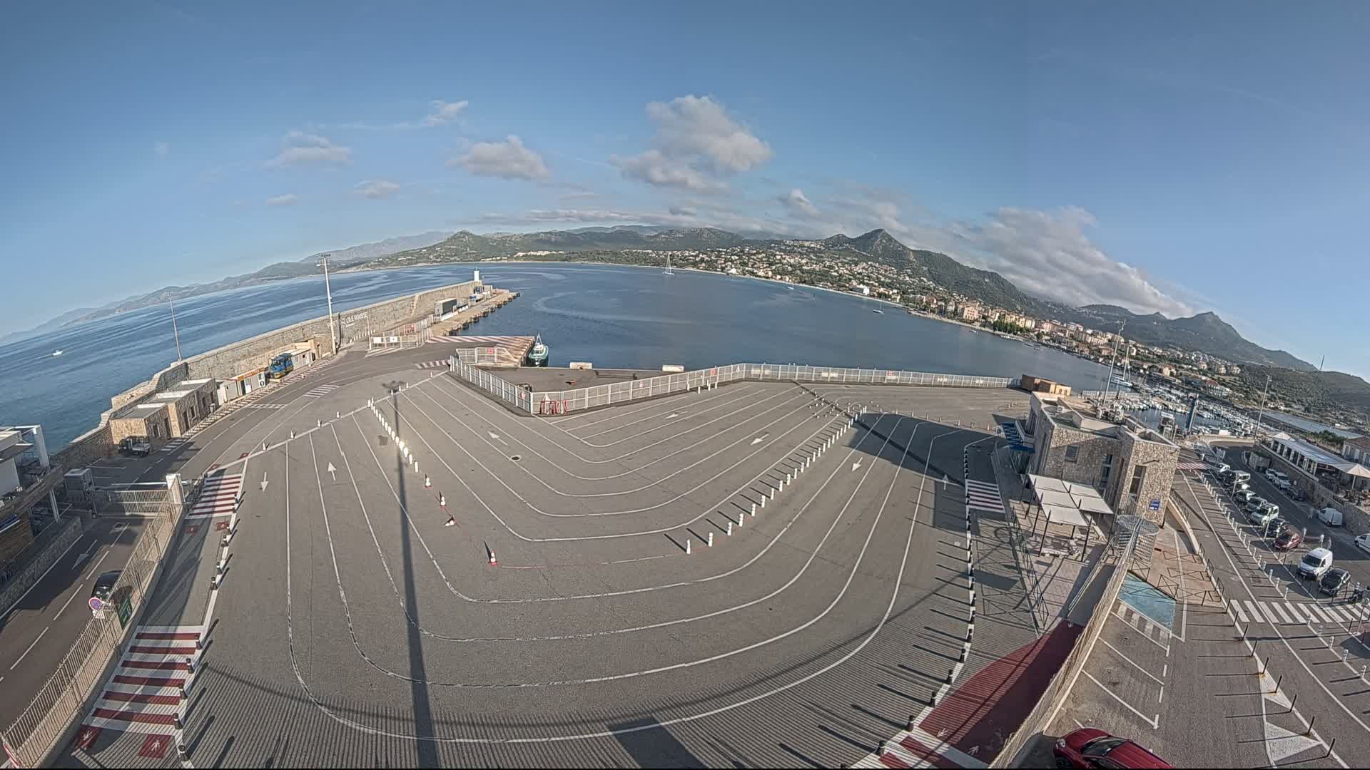 An empty ferry terminal parking lot with marked lanes overlooks a calm bay and distant mountains under a mostly clear sky.