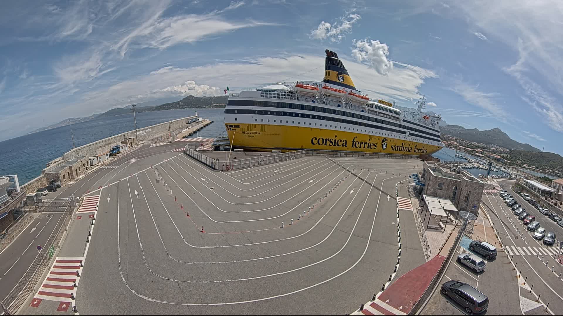 A large ferry is docked at a port on a partly sunny day, with several cars parked nearby and marked lanes guiding vehicles towards it.