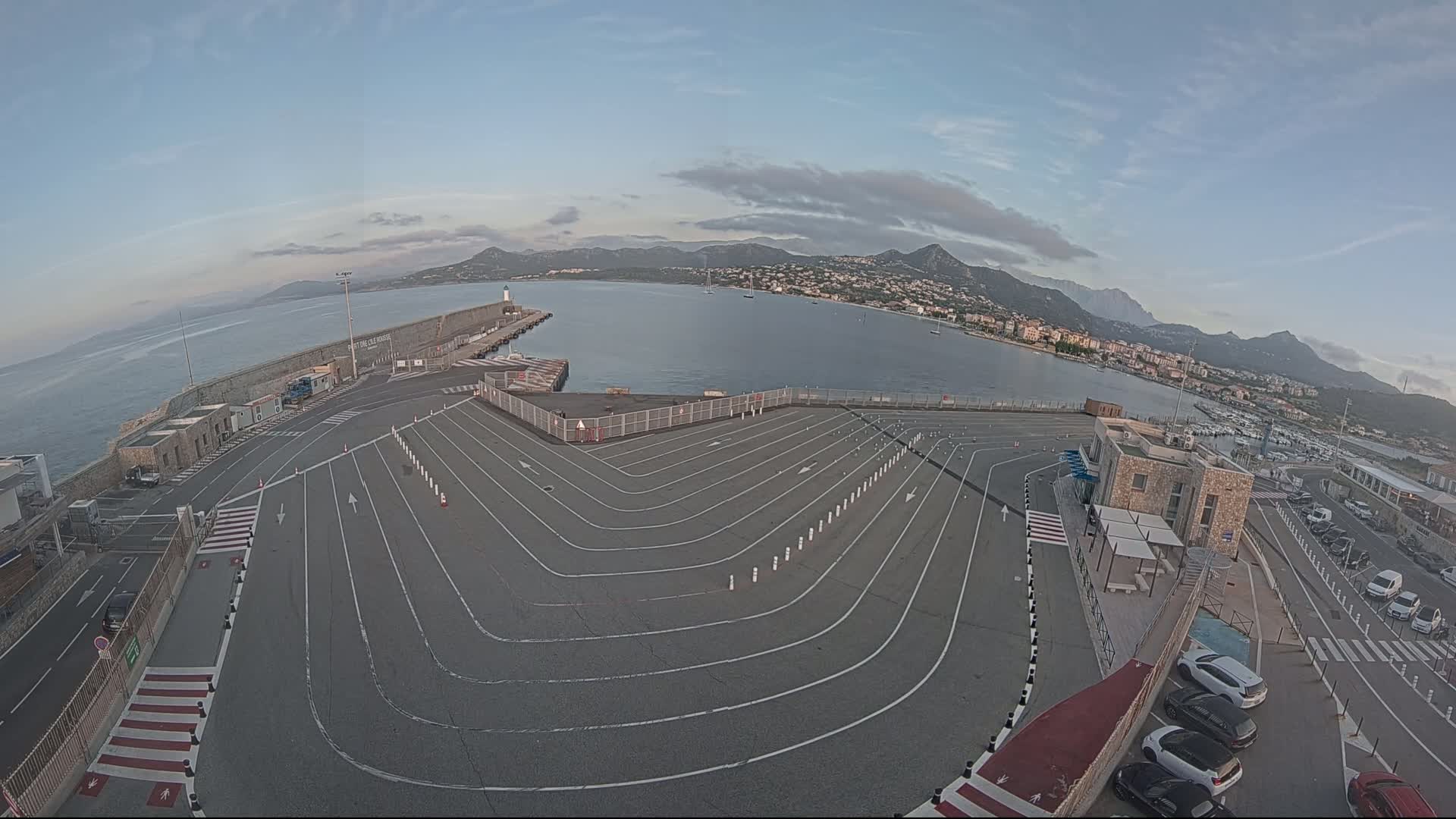 An empty vehicle staging area at a harbor overlooks calm water and distant mountains under a mostly clear sky.