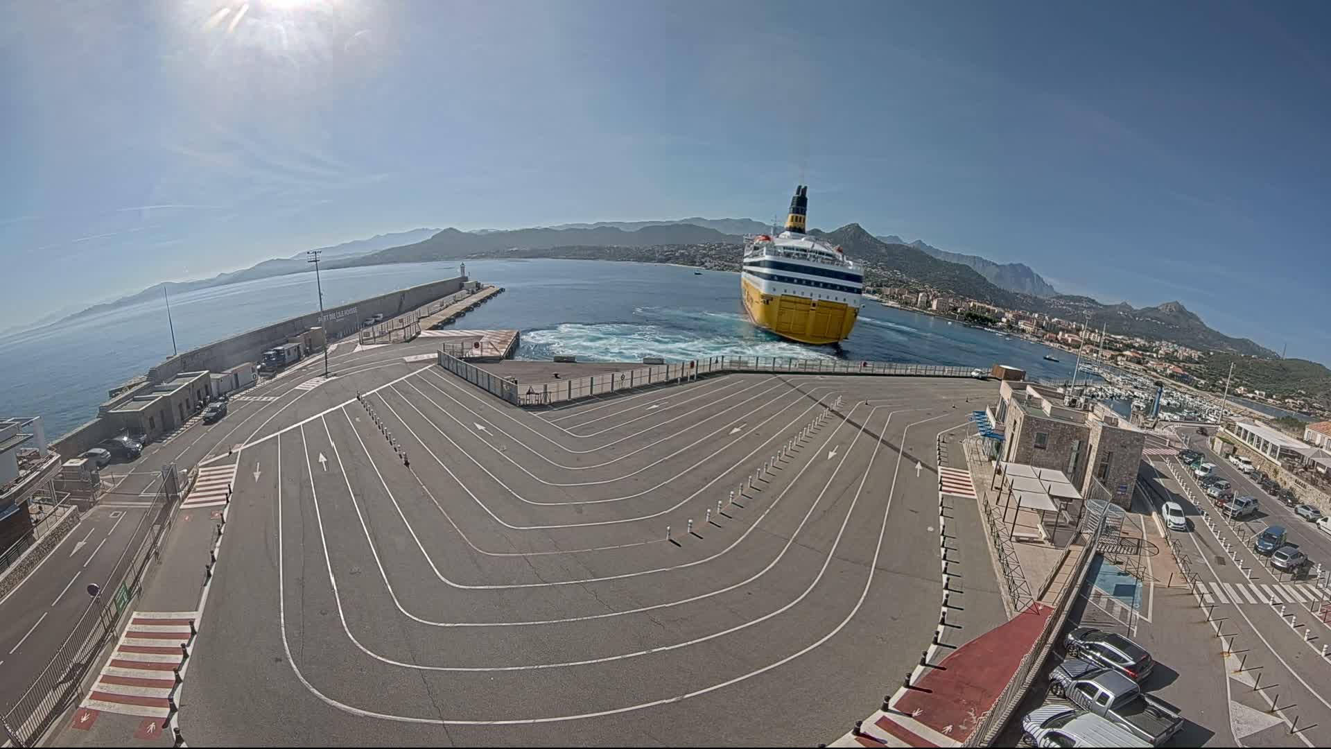 A large yellow and blue ferry is approaching a port on a sunny day, maneuvering into a designated parking area marked with white lines.