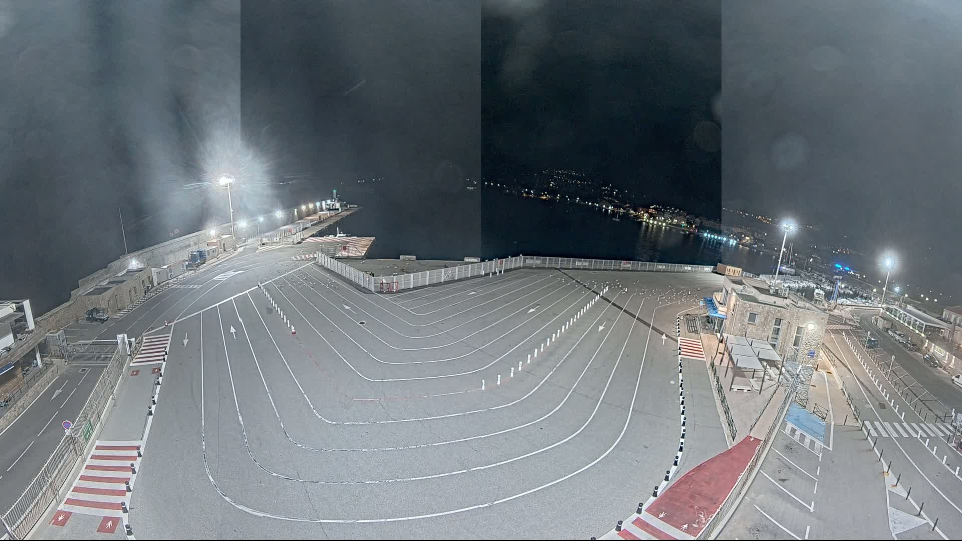 An empty, paved ferry terminal at night under a dark, cloudy sky is illuminated by artificial lights, with a calm body of water and a town visible in the distance.