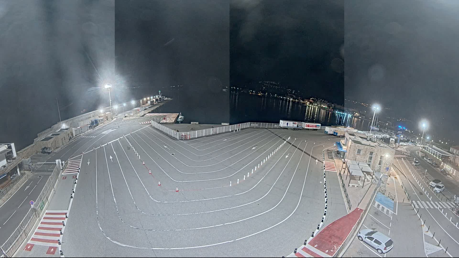 An empty, marked vehicle staging area at a harbor at night under a dark, cloudy sky.
