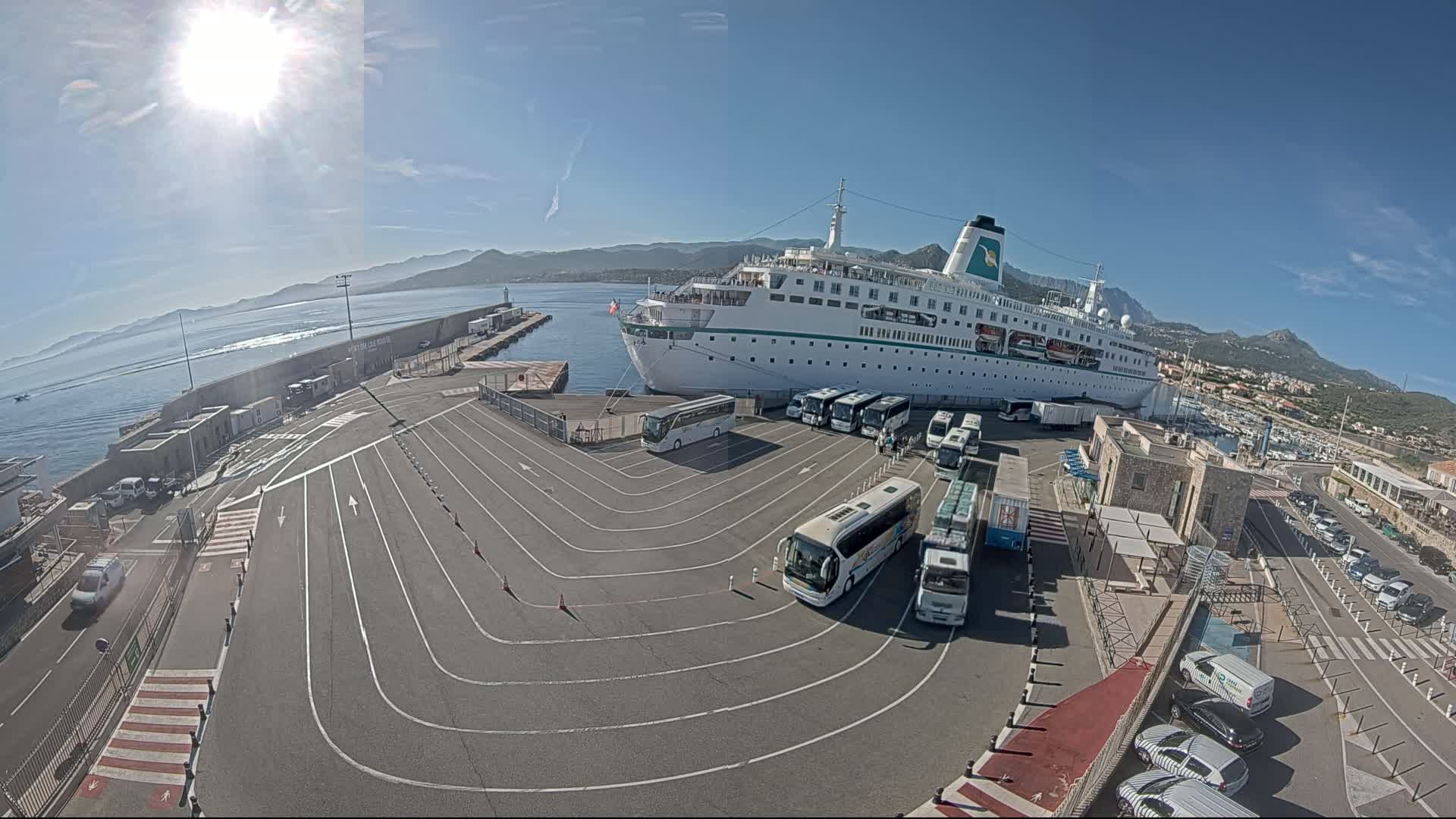 A large cruise ship is docked at a port on a sunny day, surrounded by numerous buses and cars in a paved area.