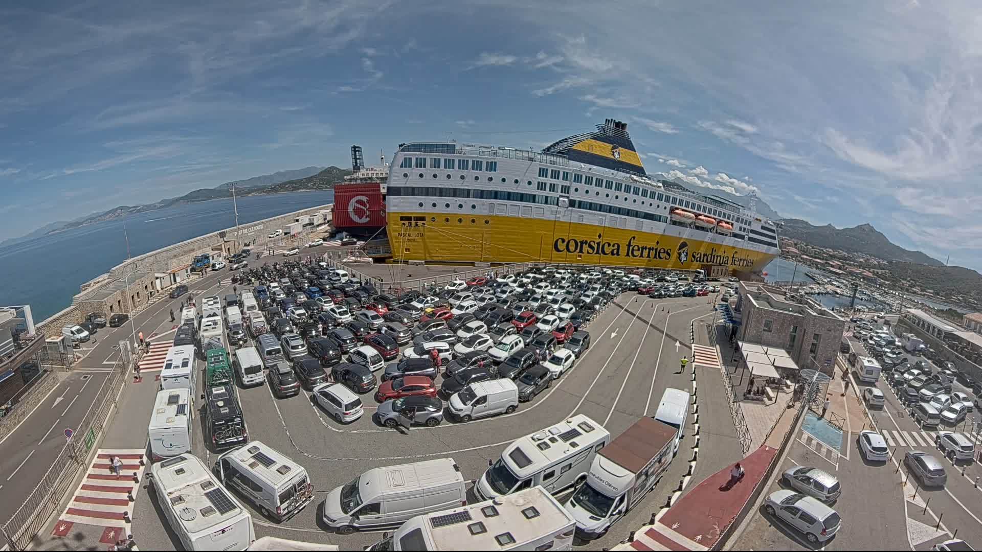 A large yellow and black ferry is docked at a port on a sunny day, surrounded by numerous cars and campervans in a large parking area.