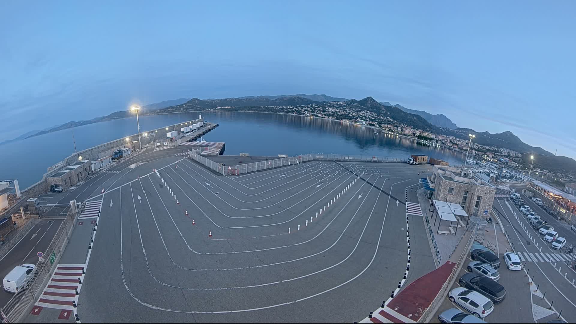 An empty, marked parking lot at a harbor overlooks calm water and mountains under a twilight sky.