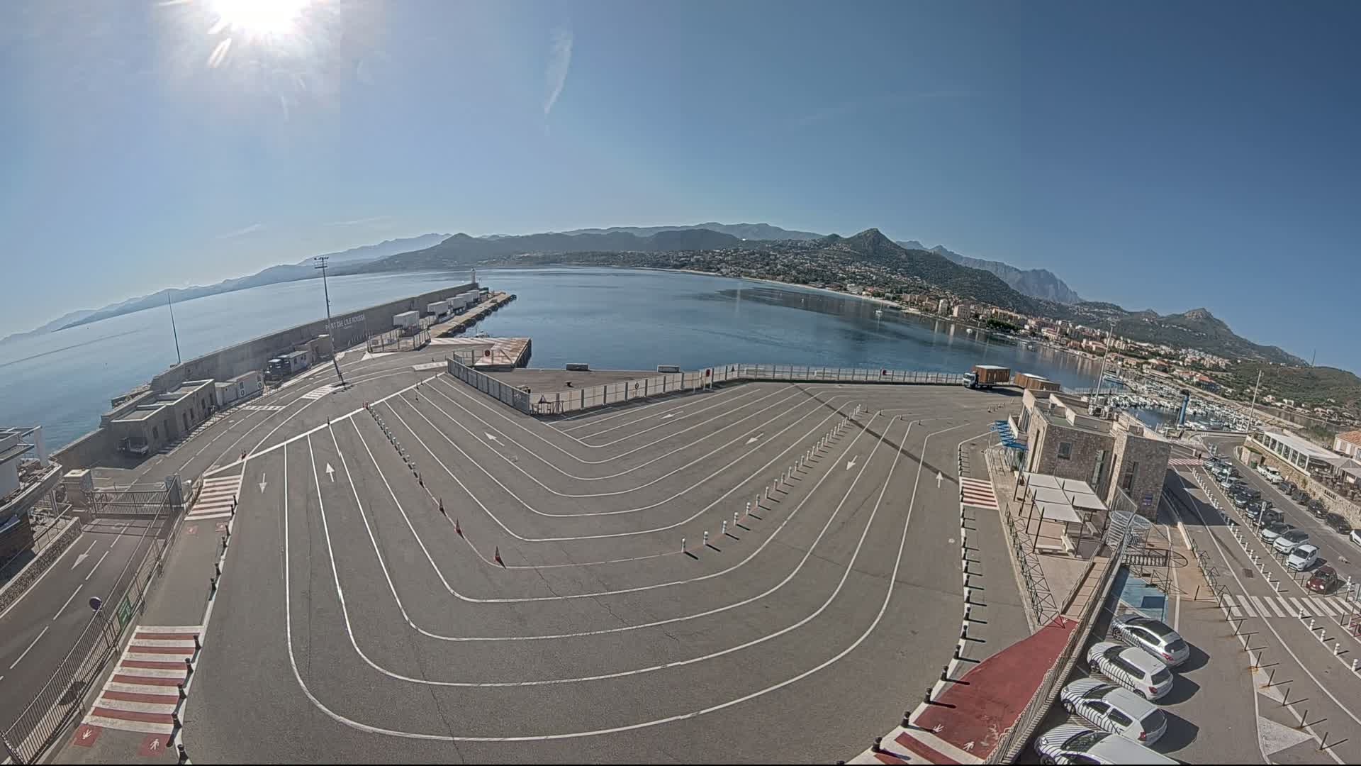 A sunny day reveals an empty ferry terminal with marked lanes curving toward a calm harbor, backed by distant mountains.