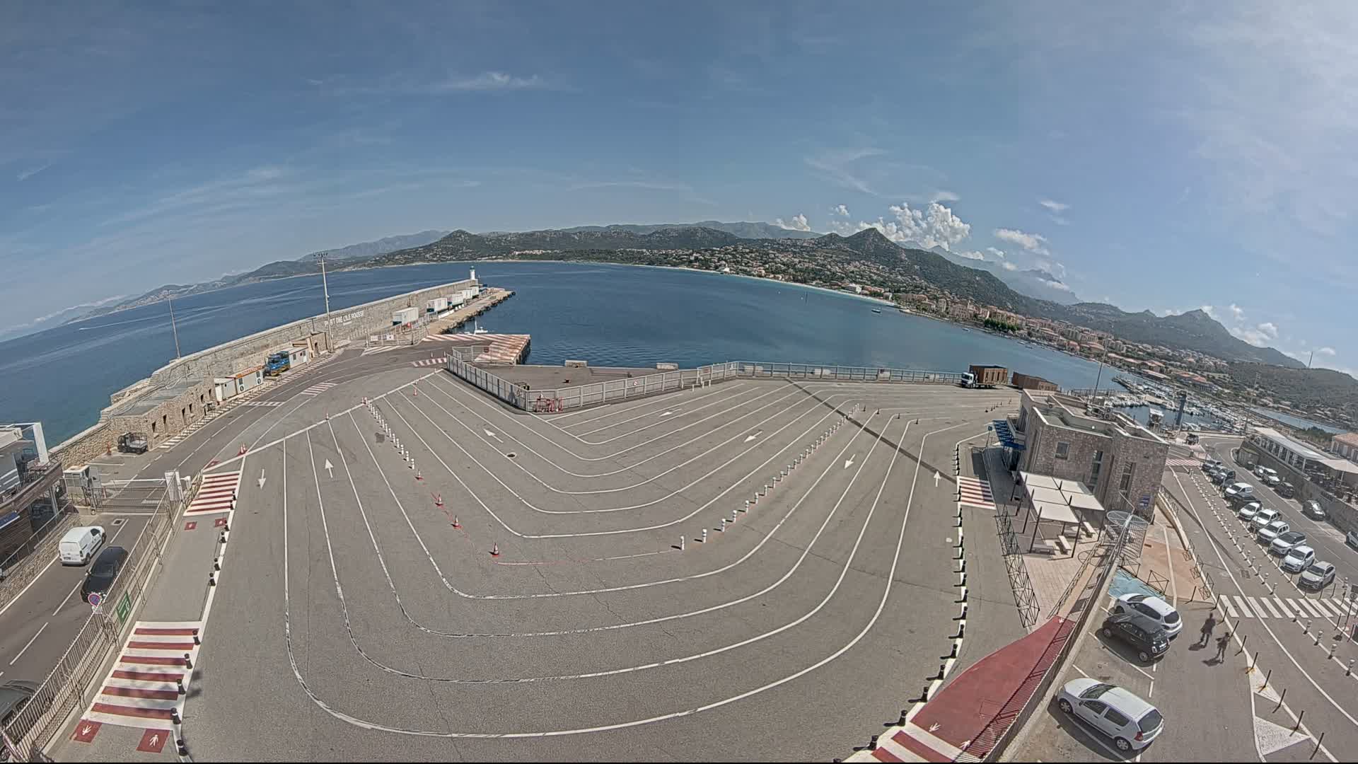 A large, empty ferry terminal with marked parking lanes overlooks a calm bay and mountainous coastline under a mostly sunny sky.