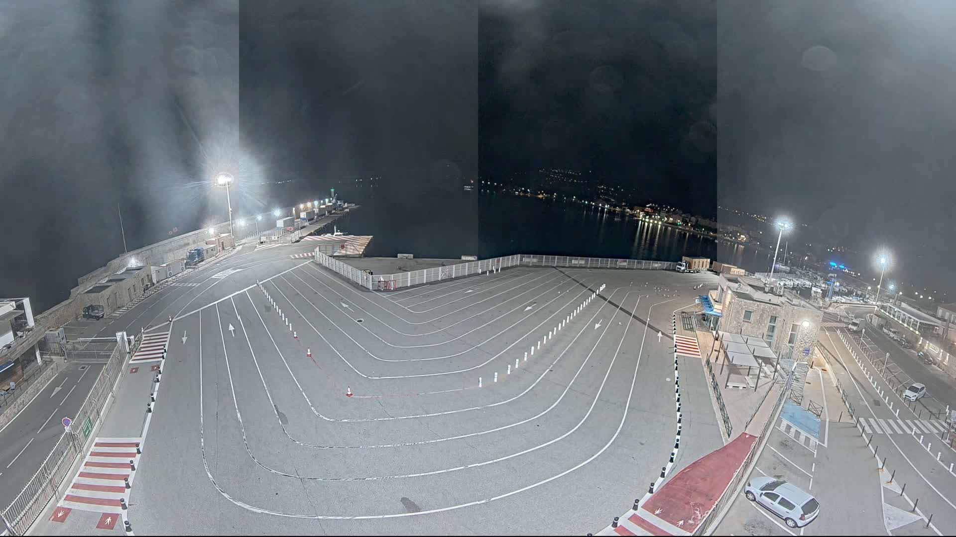 An empty, nighttime ferry terminal with marked parking lanes overlooks a calm body of water under a cloudy sky.