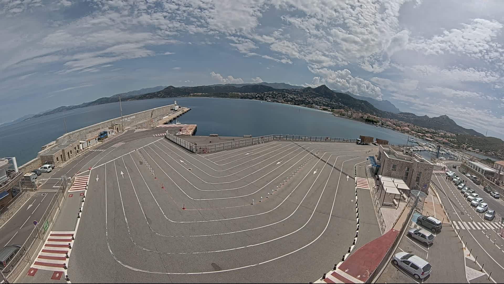 An empty ferry terminal with marked lanes overlooks a calm bay and distant mountains under a partly cloudy sky.