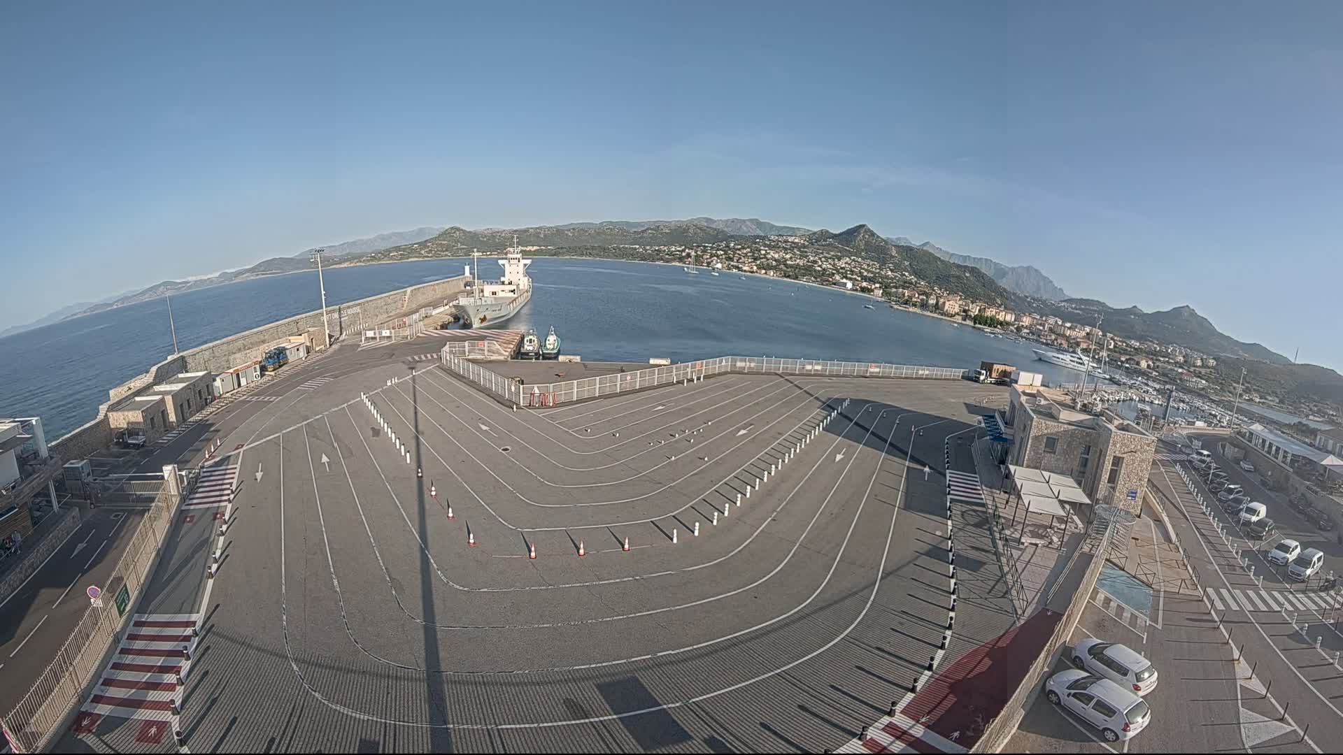 A large, empty ferry terminal with marked lanes sits beside a calm body of water, mountains in the distance under a clear blue sky.