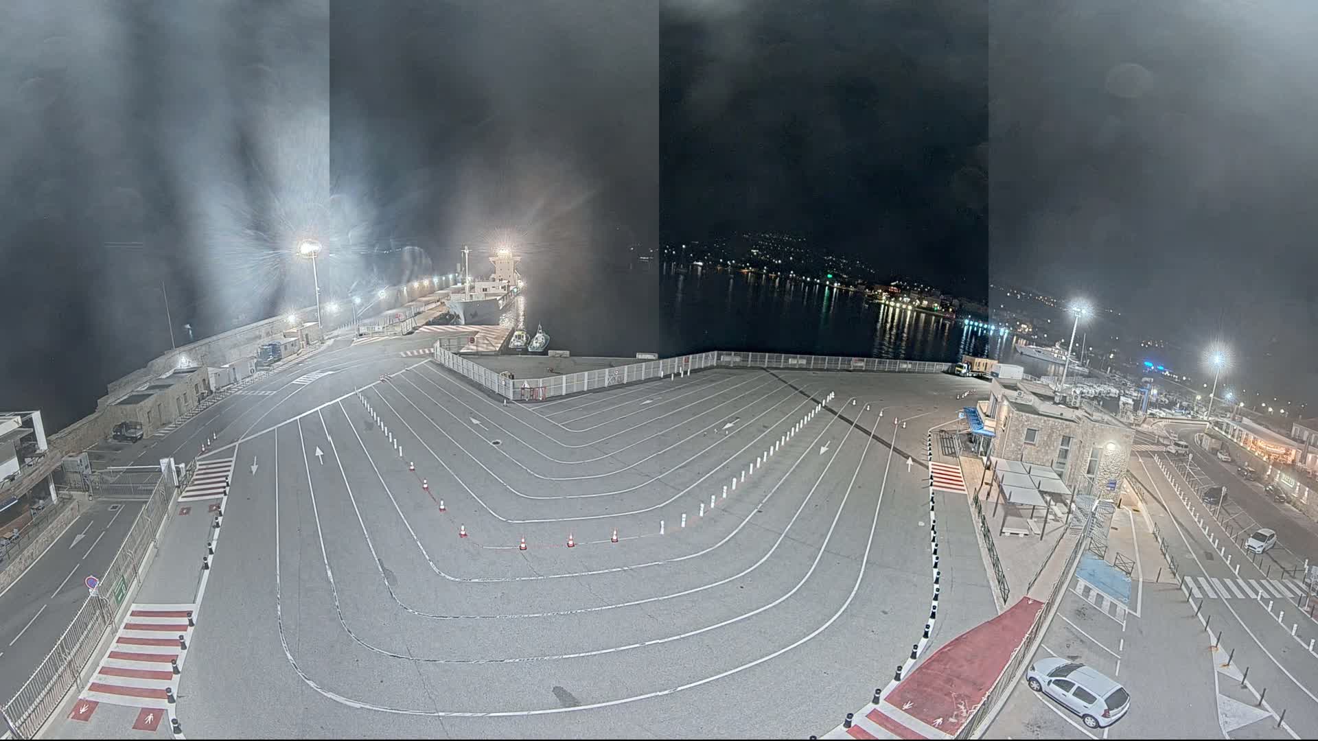 An empty, marked parking lot at a harbor at night, under a dark, cloudy sky, with a ship docked nearby and city lights visible across the water.