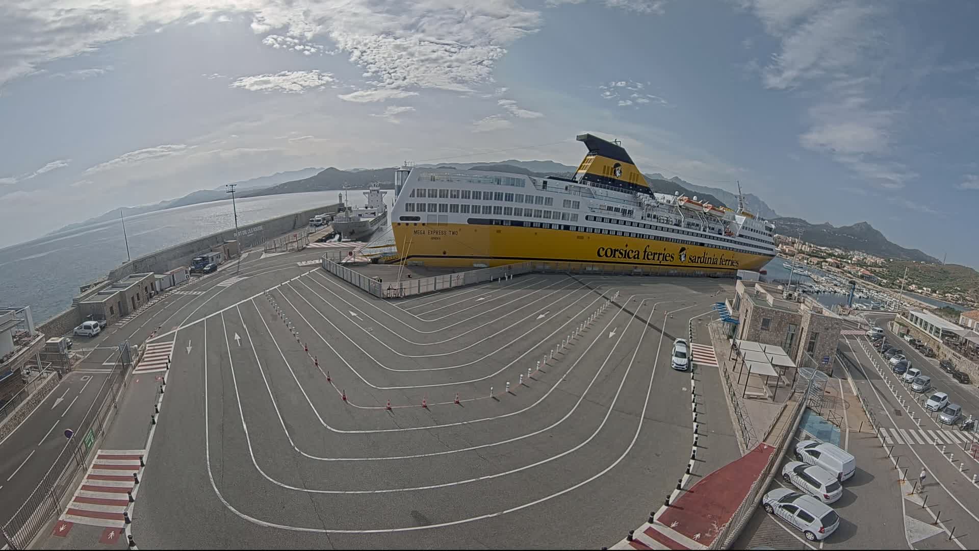 A large yellow and white ferry is docked at a port on a partly cloudy day, with several cars parked nearby and a body of water visible in the background.