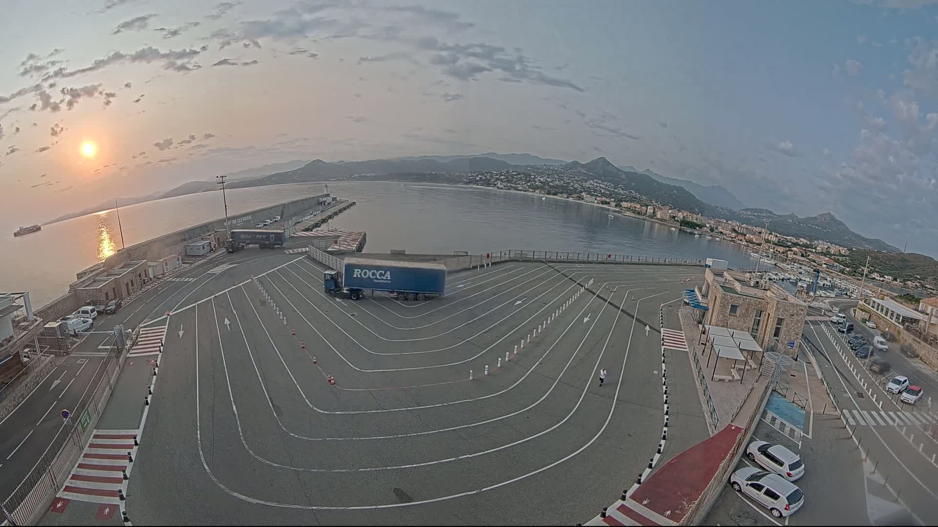 A sunrise illuminates a harbor where a semi-truck is parked in a designated area with winding traffic lanes, and cars are parked in a lot next to a small building, all overlooking calm water and distant mountains.
