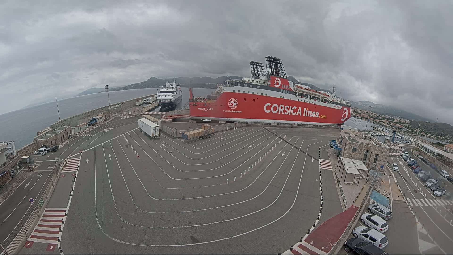 A large red ferry is docked at a port on an overcast day, with a smaller passenger ship visible in the background and several cars parked nearby.