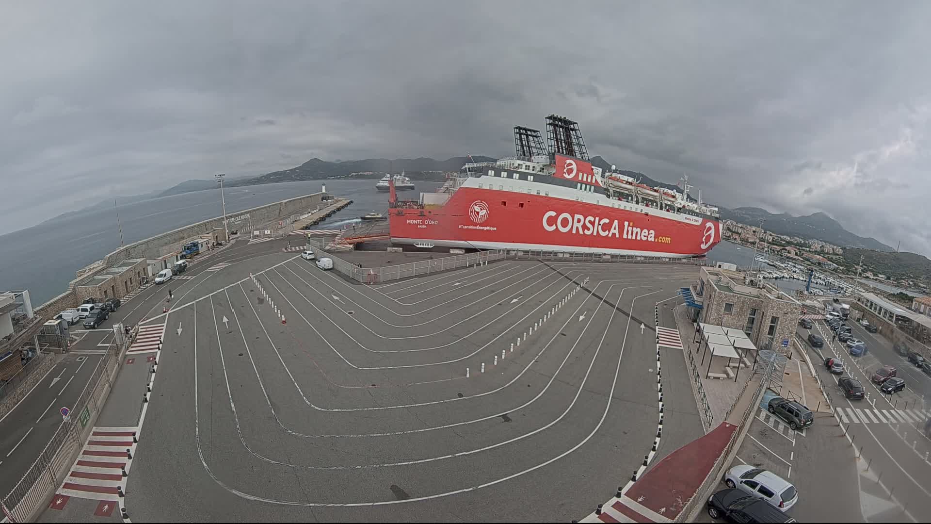 A large red ferry is docked at a port under an overcast sky, with cars parked in marked lanes nearby.