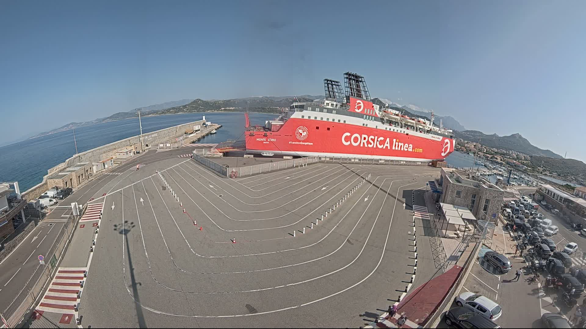 A large red ferry is docked at a port on a sunny day, with a paved area marked with driving lanes leading to it.
