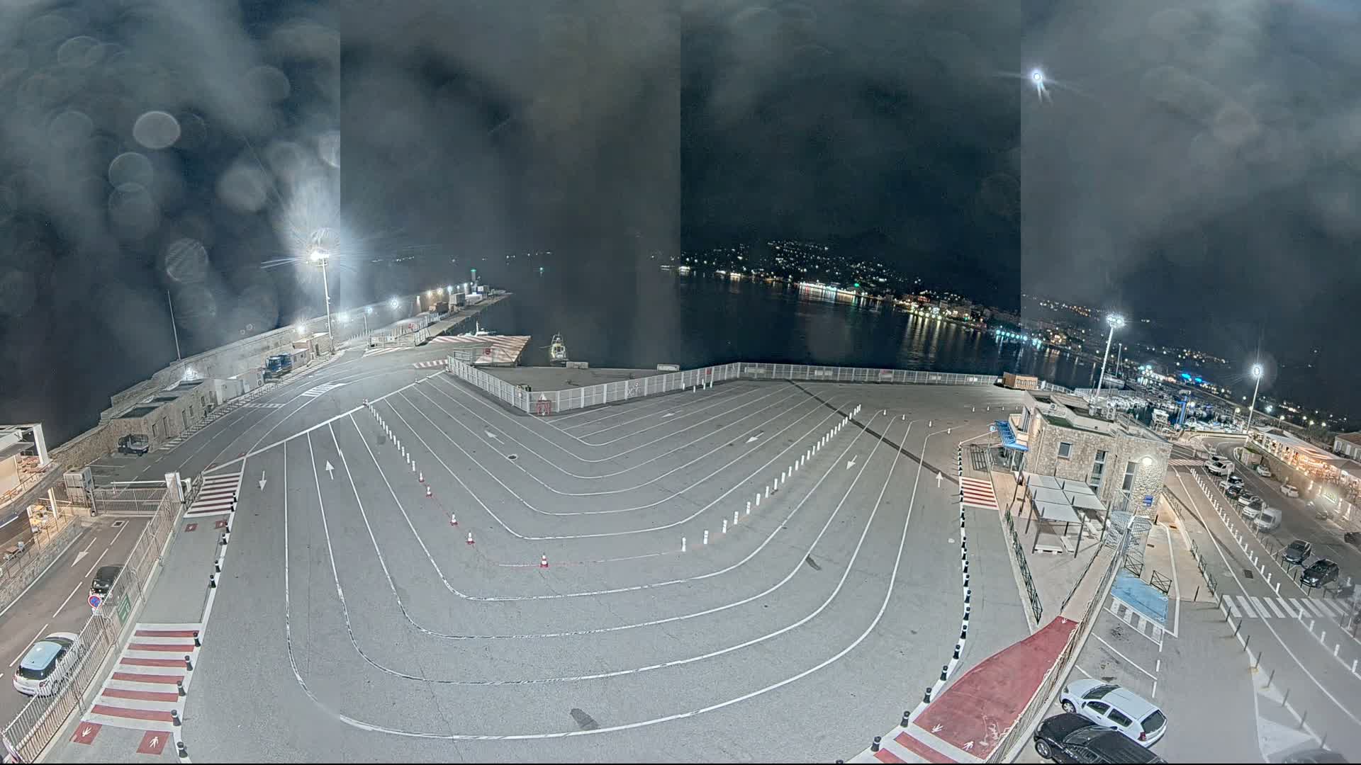 An empty, marked parking lot at a harbor at night under a cloudy sky is illuminated by lights.