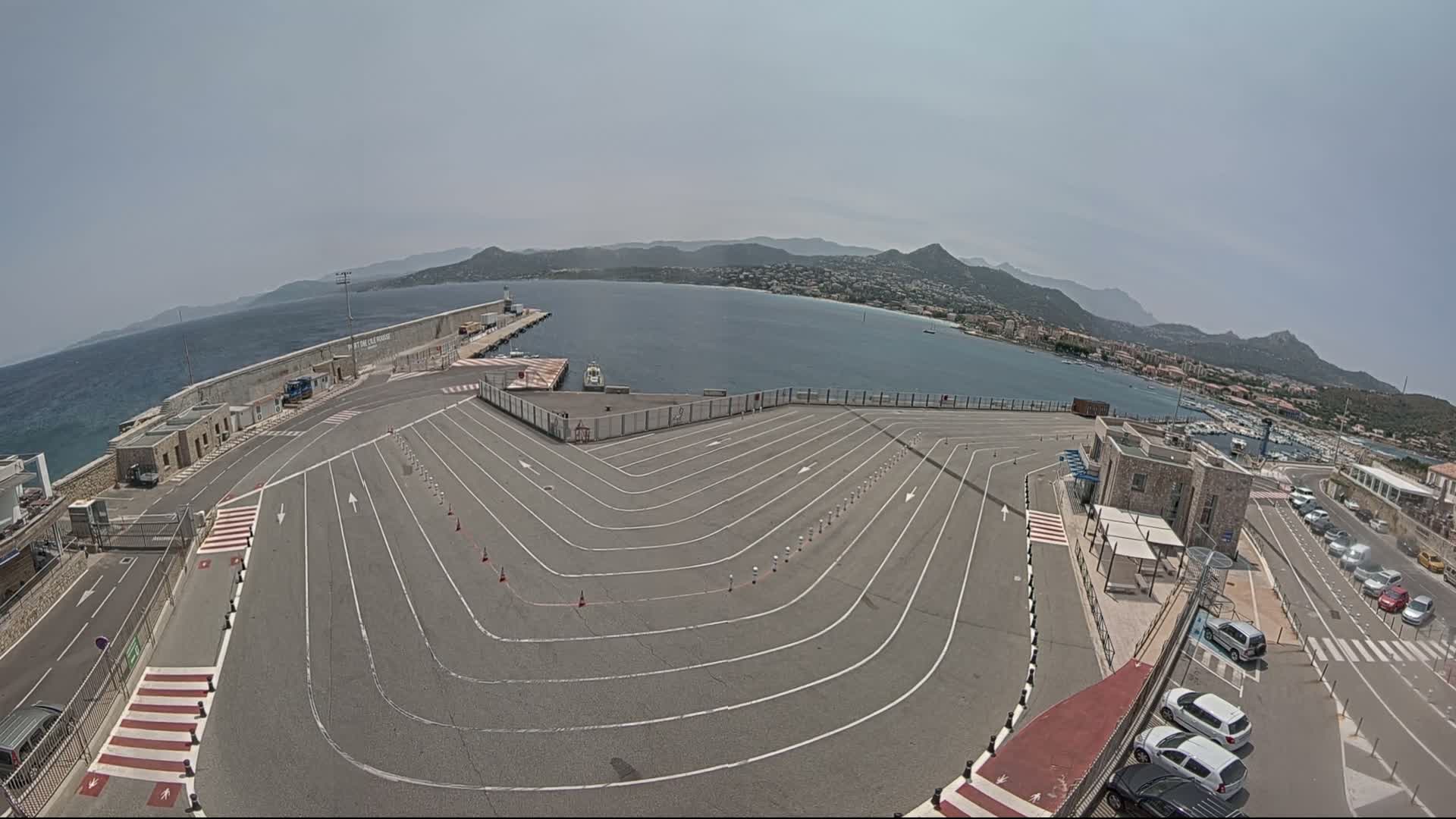 An empty, marked vehicle staging area overlooks a calm bay and distant mountains under a clear sky.