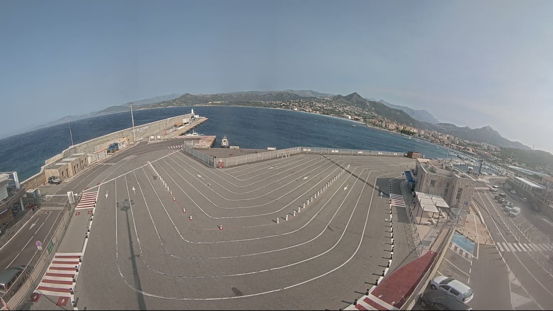 A large, empty paved area with marked lanes curves toward a harbor, overlooking a calm sea and distant mountains under a clear sky.