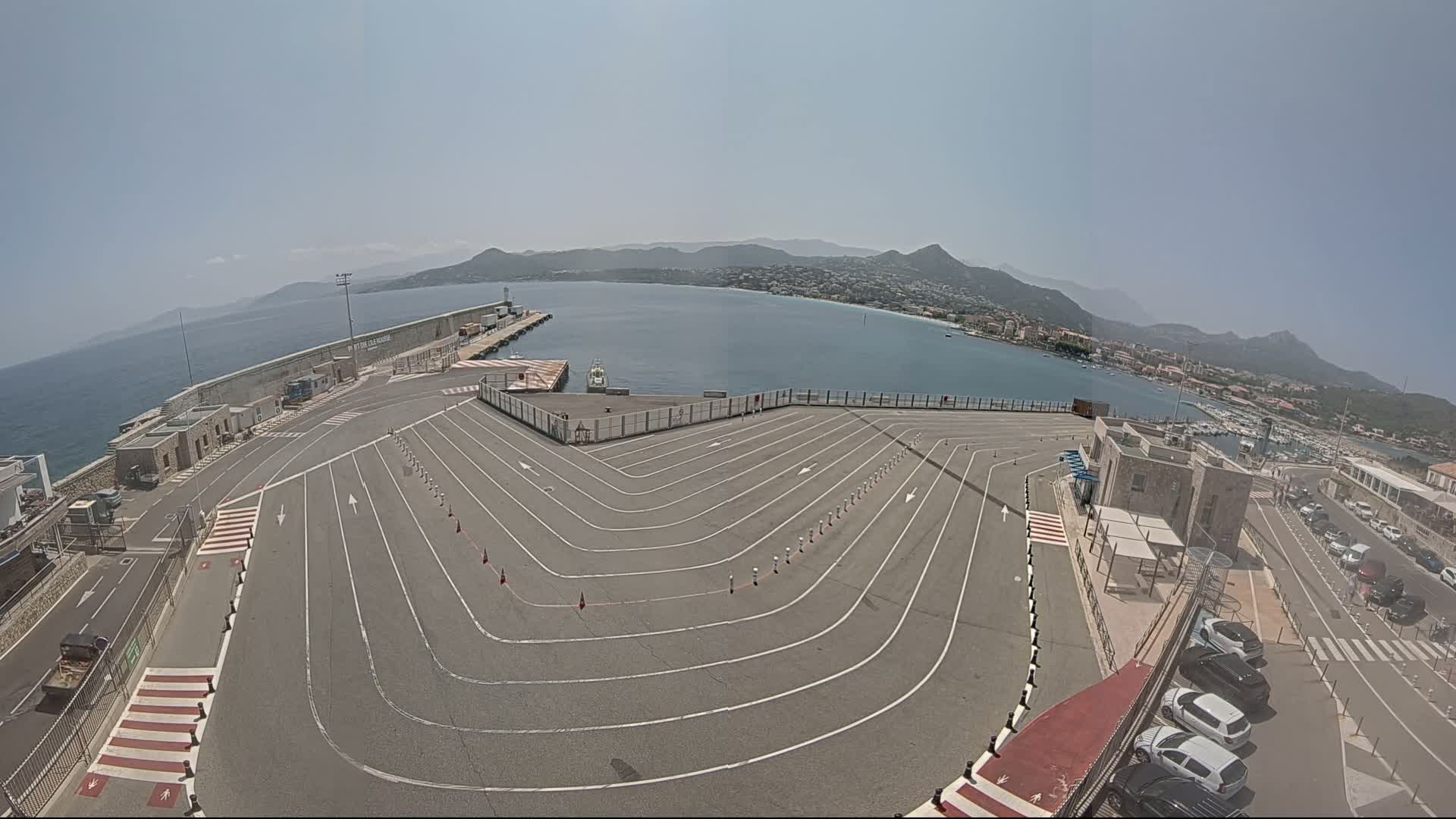 An empty ferry terminal with marked lanes curves toward a calm body of water, backed by mountains under a clear sky.