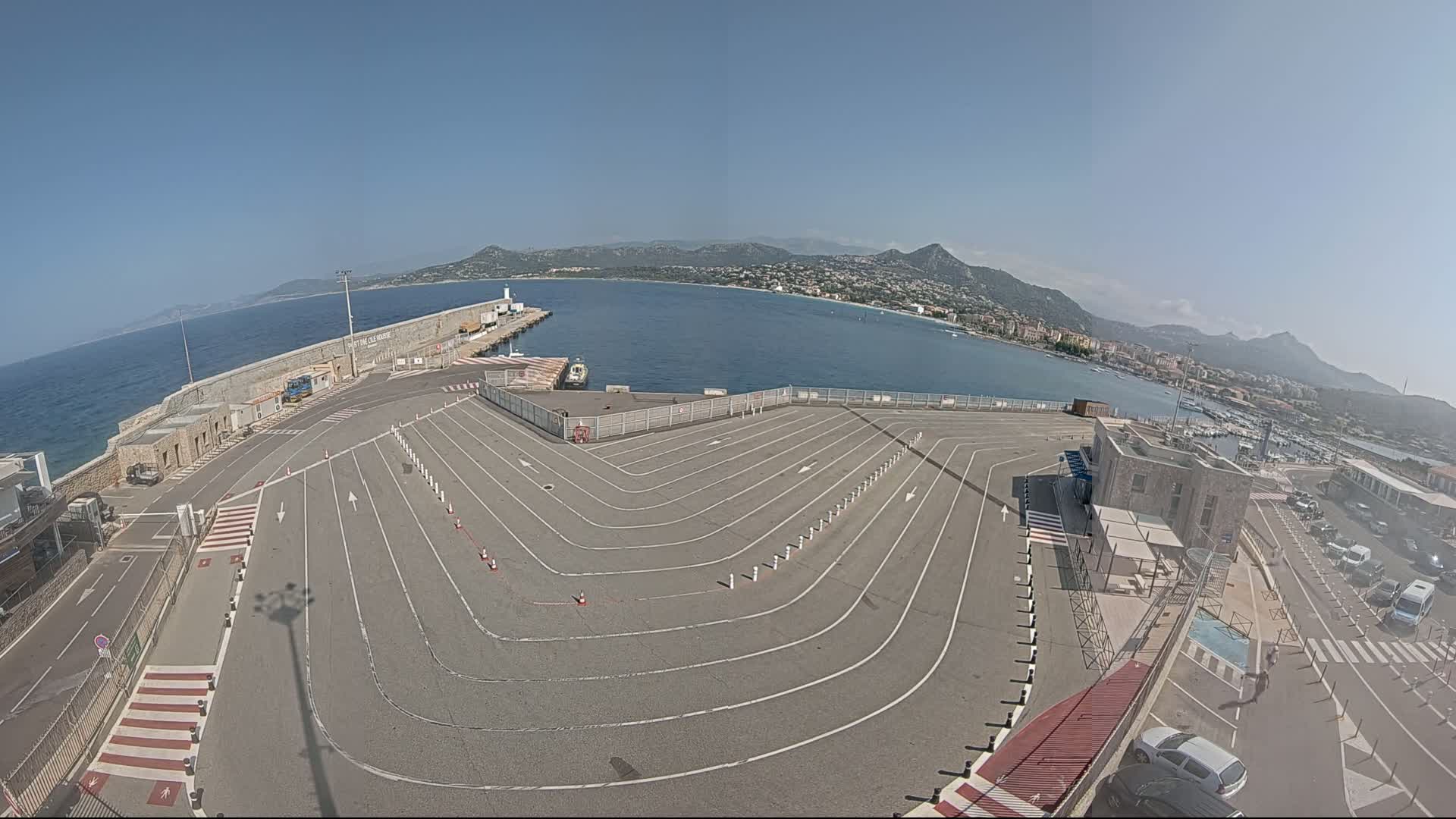 An aerial view of a port on a sunny day shows a large, empty vehicle staging area with marked lanes curving toward a body of water with a distant mountainous coastline.