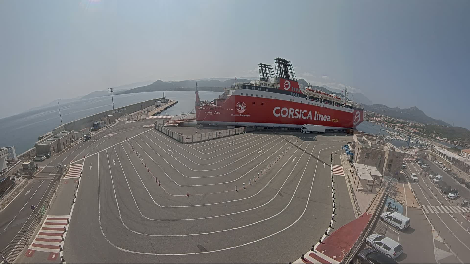 A large red ferry is docked at a port on a sunny day, with numerous cars parked nearby and a clear view of the calm ocean and distant mountains.