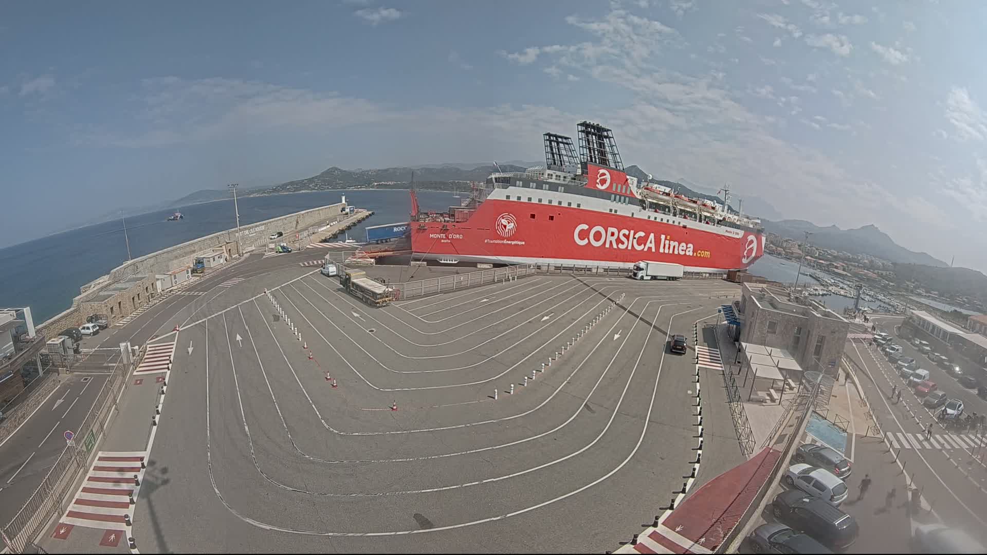 A large red ferry is docked at a port on a sunny day, with cars parked in designated lanes leading to the vessel.
